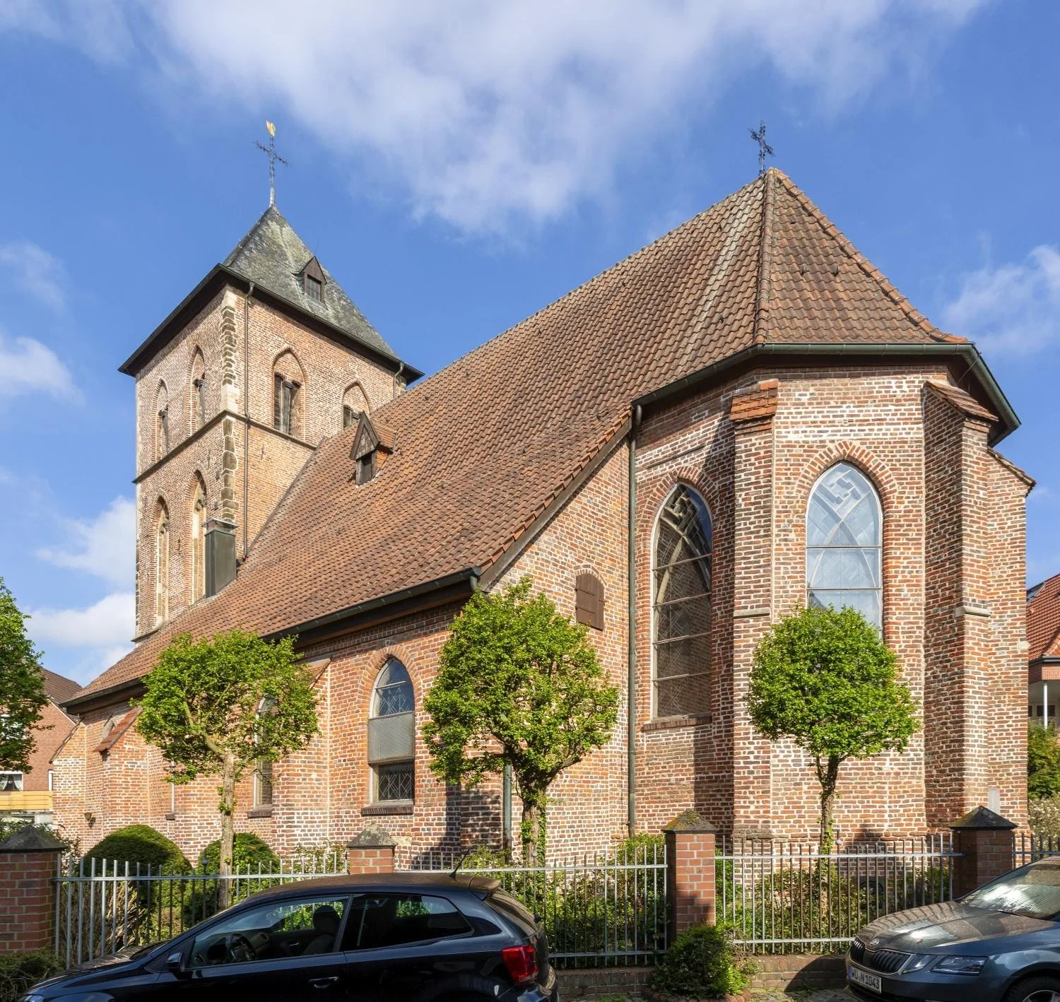 Eine historische Backsteinkirche mit einem hohen Glockenturm und roten Ziegeldächern, umgeben von grünen Bäumen und einem Zaun, bei sonnigem Himmel.