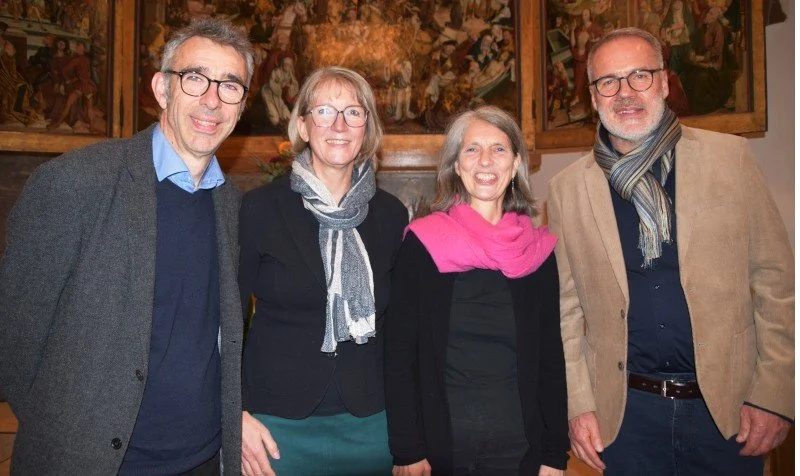 Vier Erwachsene stehen in einem Raum vor einer Wand mit Gemälden. Sie lachen und tragen formelle Kleidung, einige mit Schals.