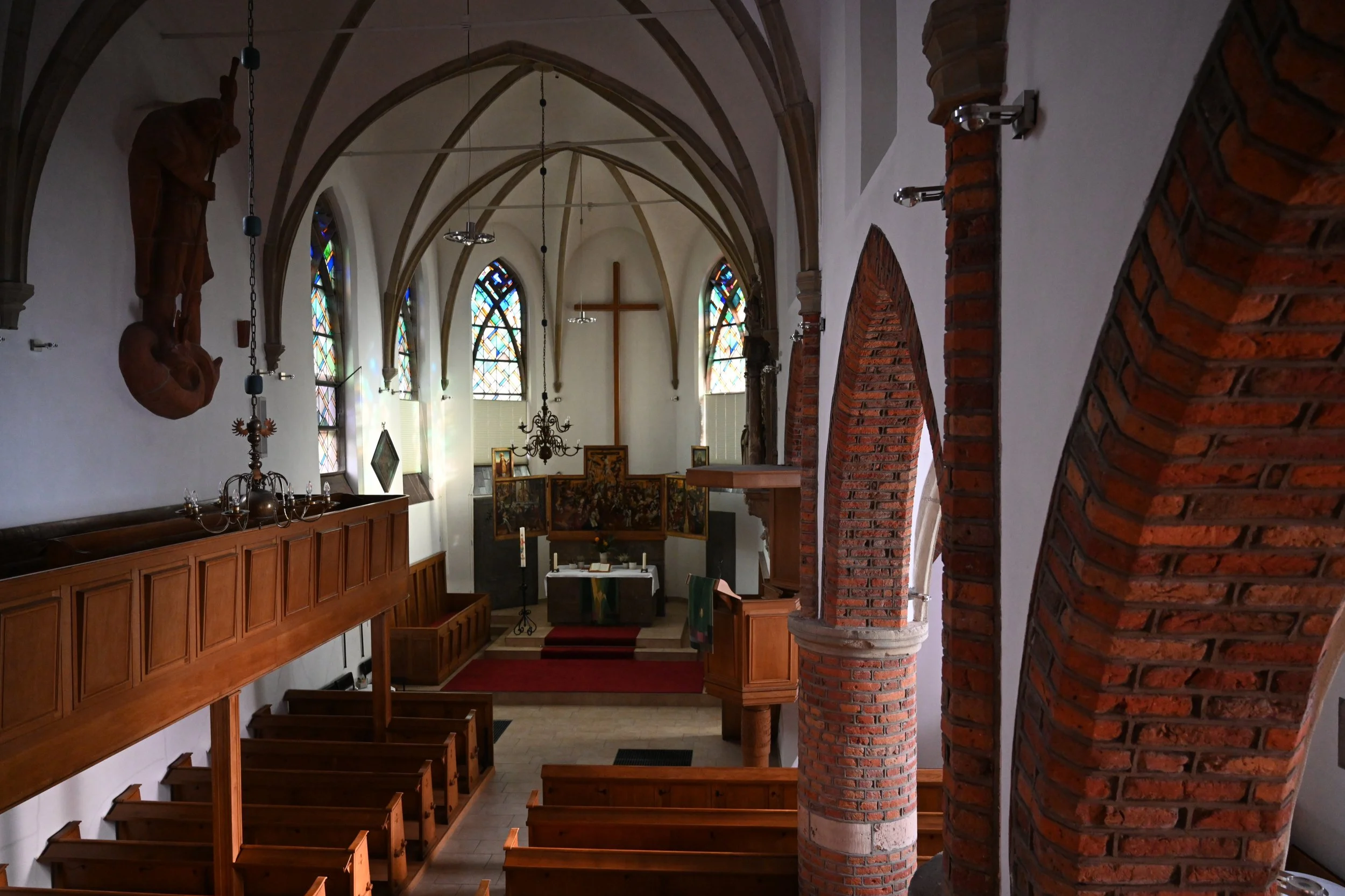 Innenansicht einer Kirche mit Holzbänken, Altar, bunten Fenstern und einem Kreuz an der Wand