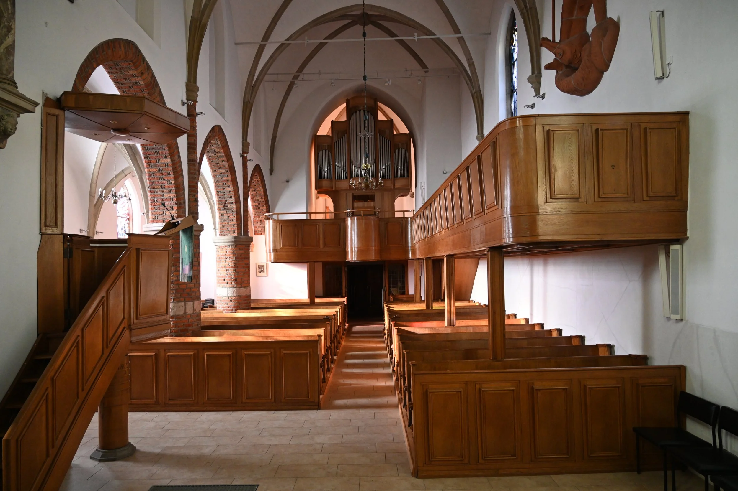 Innenansicht einer Kirche mit Holzgalerie, Buntglasfenstern und einer Orgel im Hintergrund.