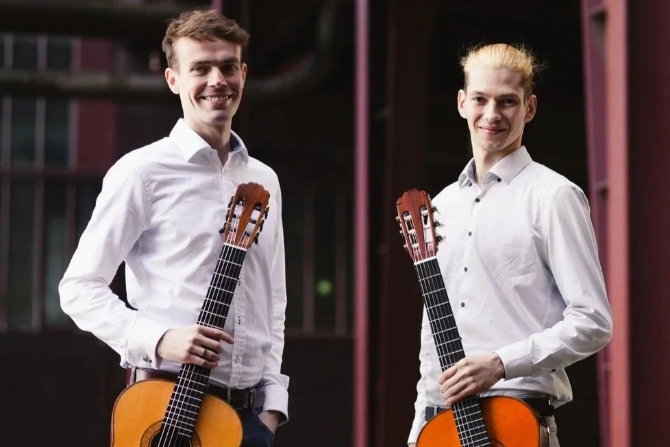 Zwei junge Männer stehen mit Gitarren vor einem dunklen Gebäude, tragen weiße Hemden und lächeln.