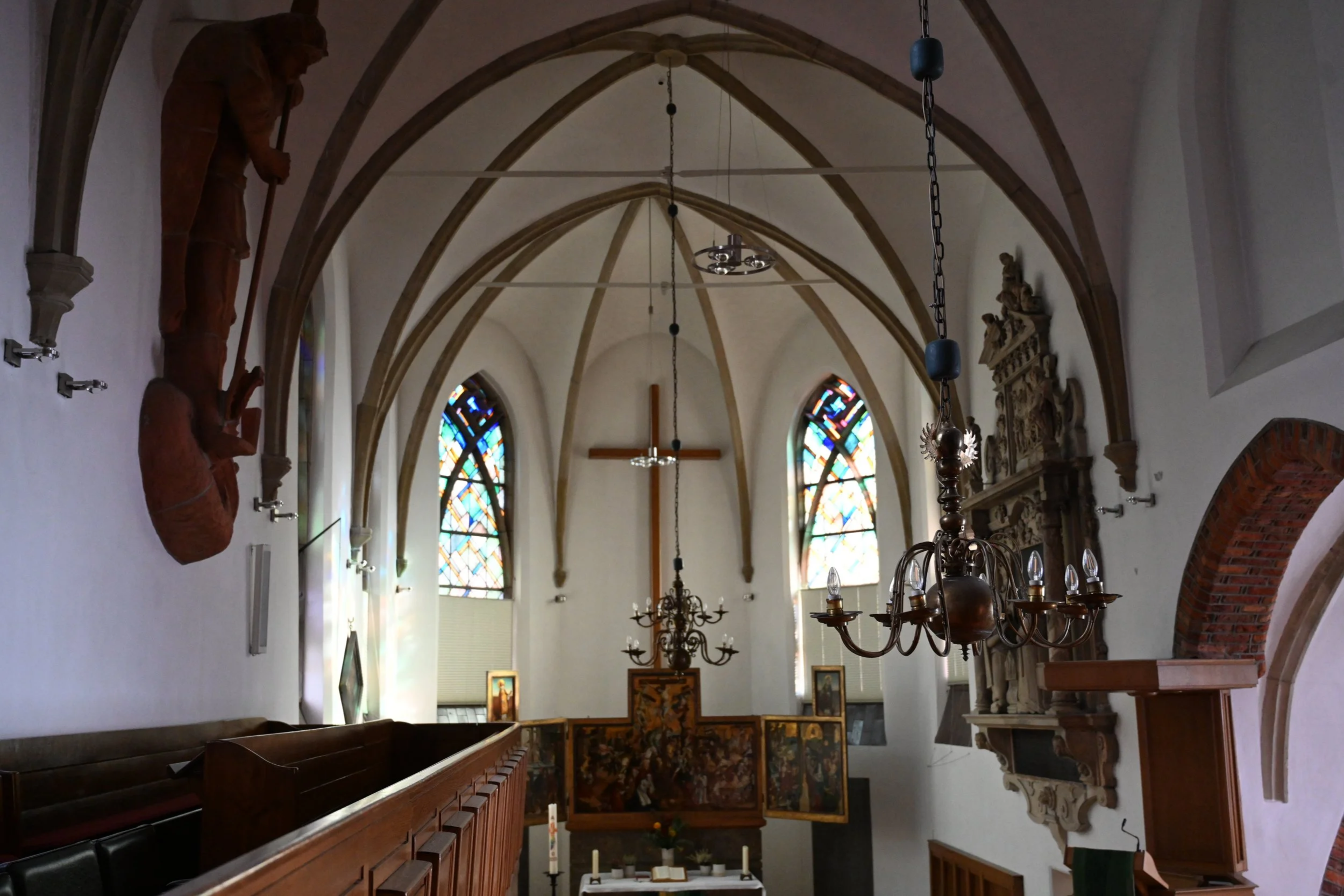 Innenansicht einer Kirche mit bunten Kirchenfenstern, einem großen Holzkreuz hinter dem Altar, Kronleuchtern, Wandreliefs und Skulpturen.