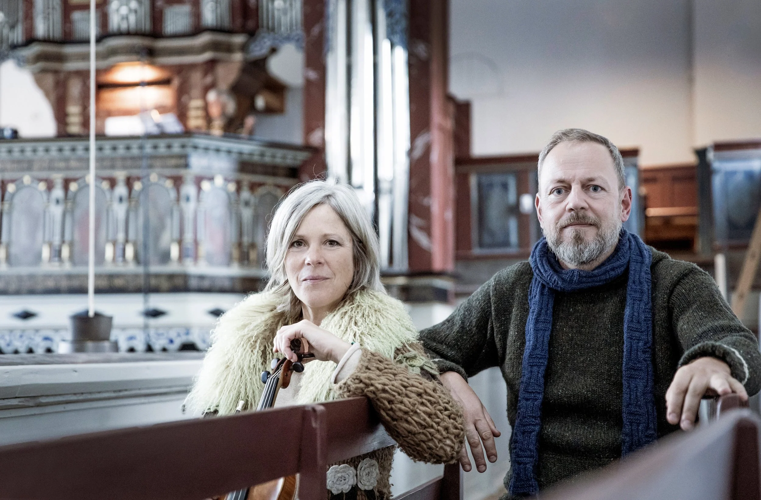 Ein Mann und eine Frau sitzen in einer Holzkirche, die Frau hält eine Geige, beide schauen in die Kamera.