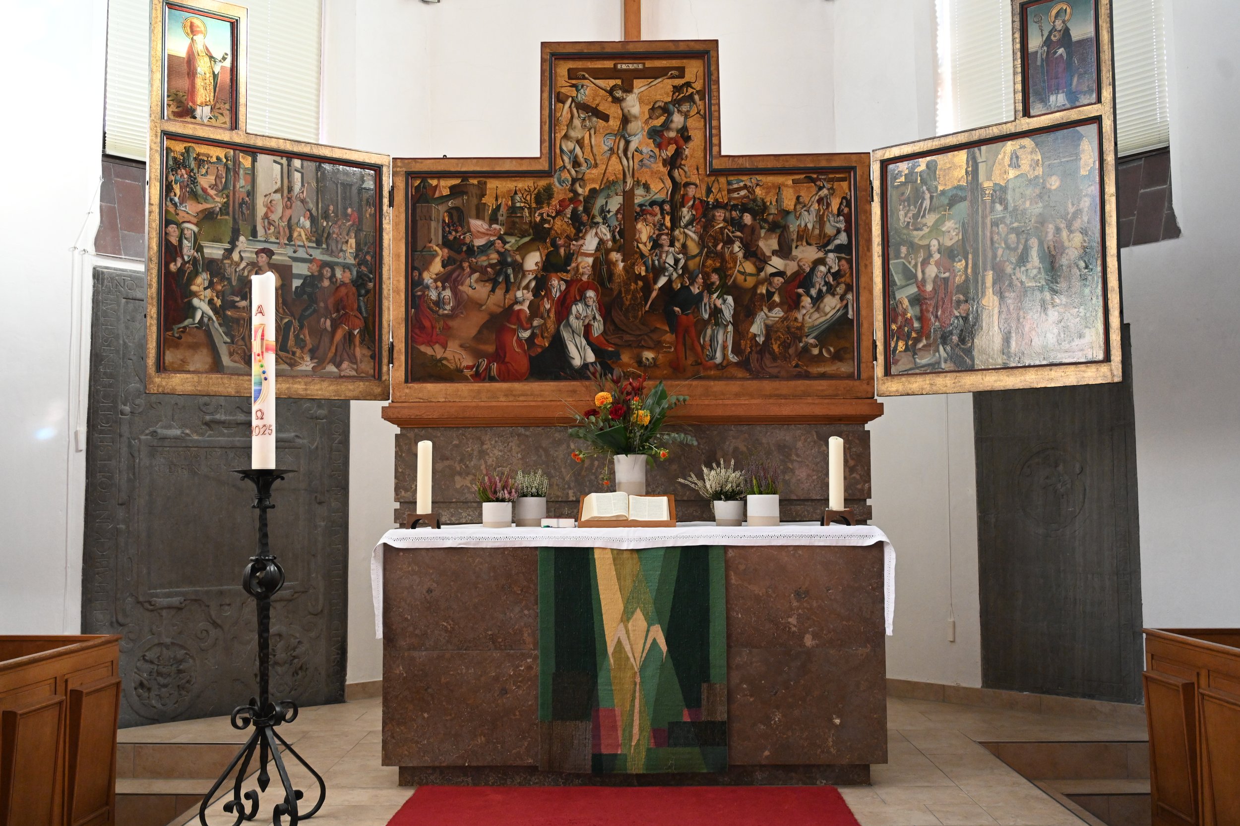 Altar in einer Kirche mit einem großen Triptychon, das die Kreuzigung Jesu zeigt. Der Altar ist mit Blumen, Kerzen und einem offenen Buch dekoriert.