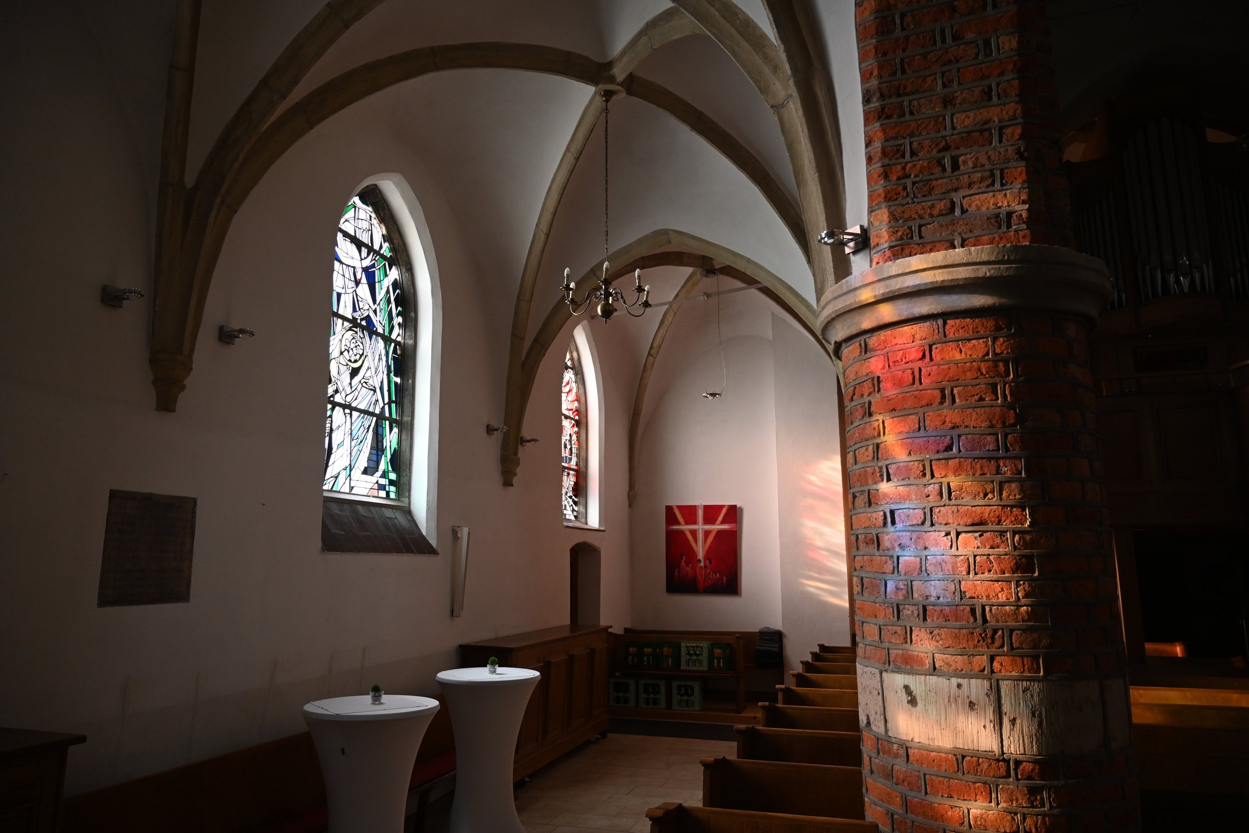 Innenansicht einer kleinen Kirche mit bunten Fenstern, einer roten Kreuz-Ikone an der Wand, Holzbankreihen und einem großen roten Ziegelmauerpfeiler.