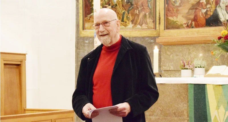 Ein älterer Mann mit Brille, rotem Hemd und schwarzer Jacke steht vor einem Altar in einer Kirche, hält Papiere in den Händen und lächelt.