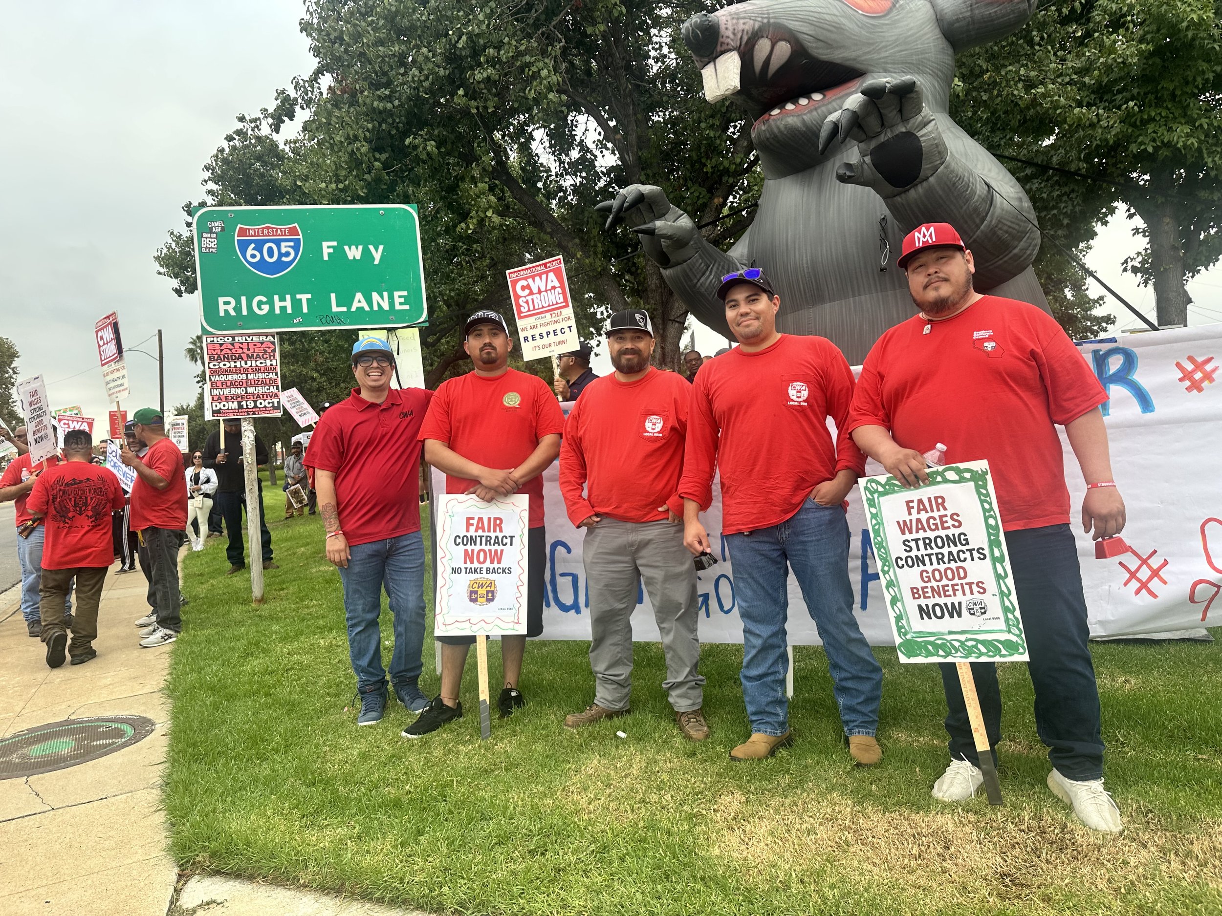 A group of five people wearing red shirts and holding protest signs standing in front of a large inflatable rat at a protest. There are more protesters and signs in the background along with a green highway sign for freeway 605.