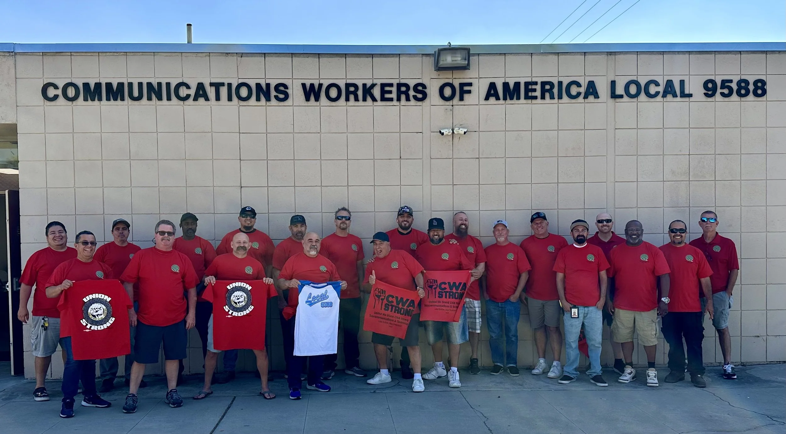 Group of union workers standing in front of a building with a sign that reads 'Communications Workers of America, Local 9588.' They are wearing red shirts, some holding banners and shirts with union logos.