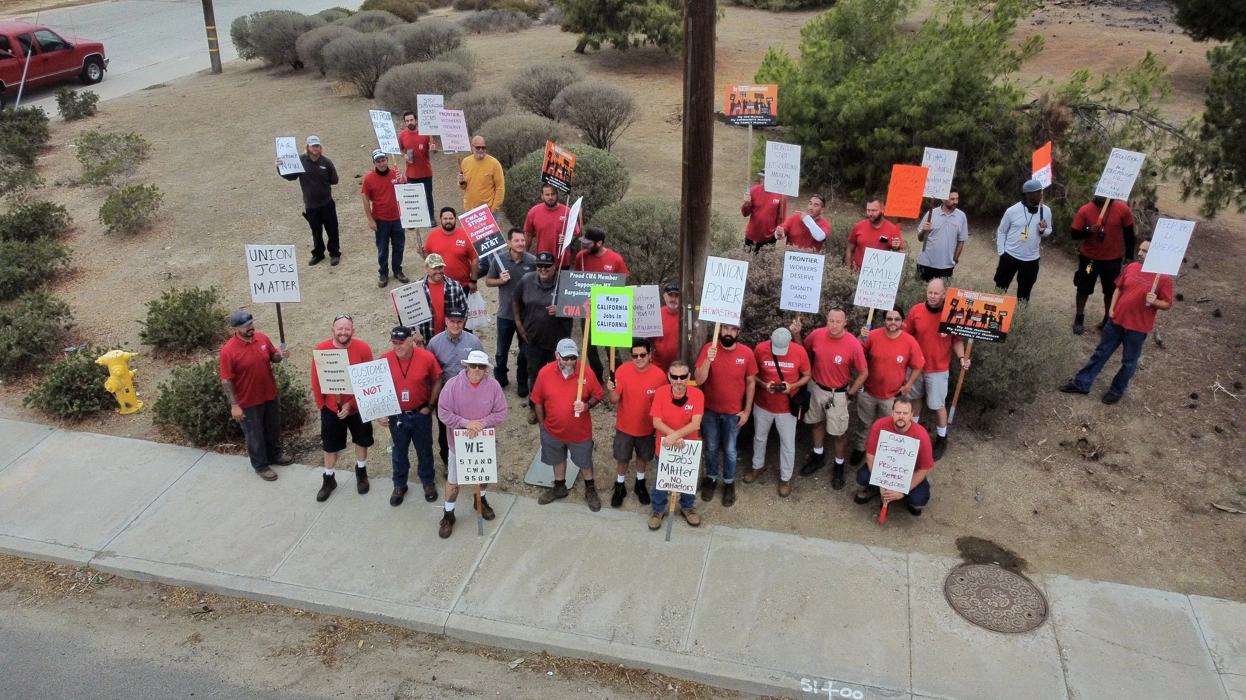 A group of protesters holding signs and banners, some wearing red shirts, gathered on a sidewalk near bushes and trees, protesting for workers' rights and union issues.