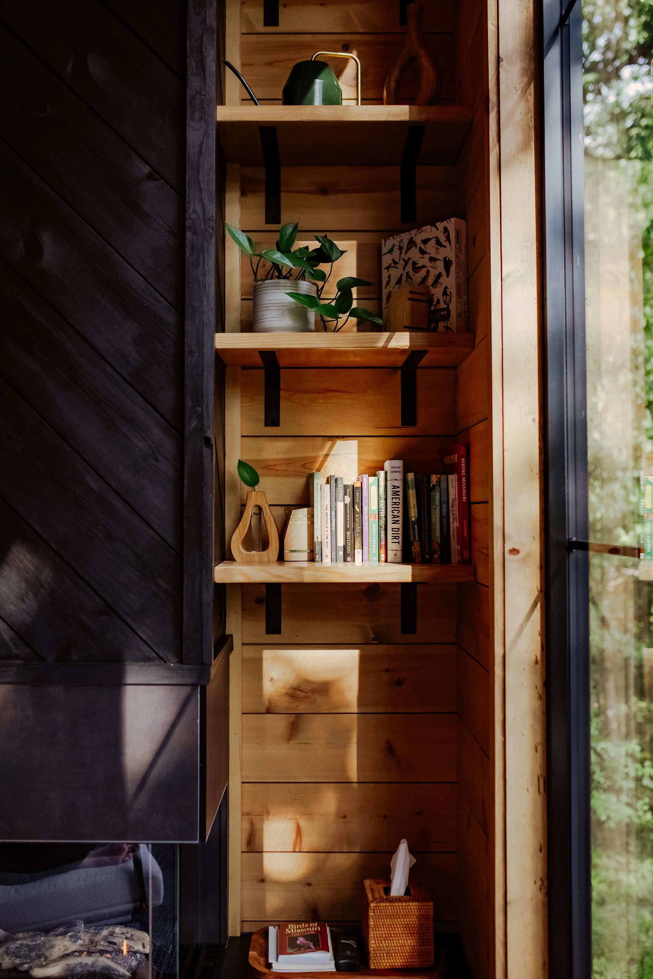 Wooden bookshelf with a potted plant, a collection of books, and decorative items, next to a large window showing trees outside.