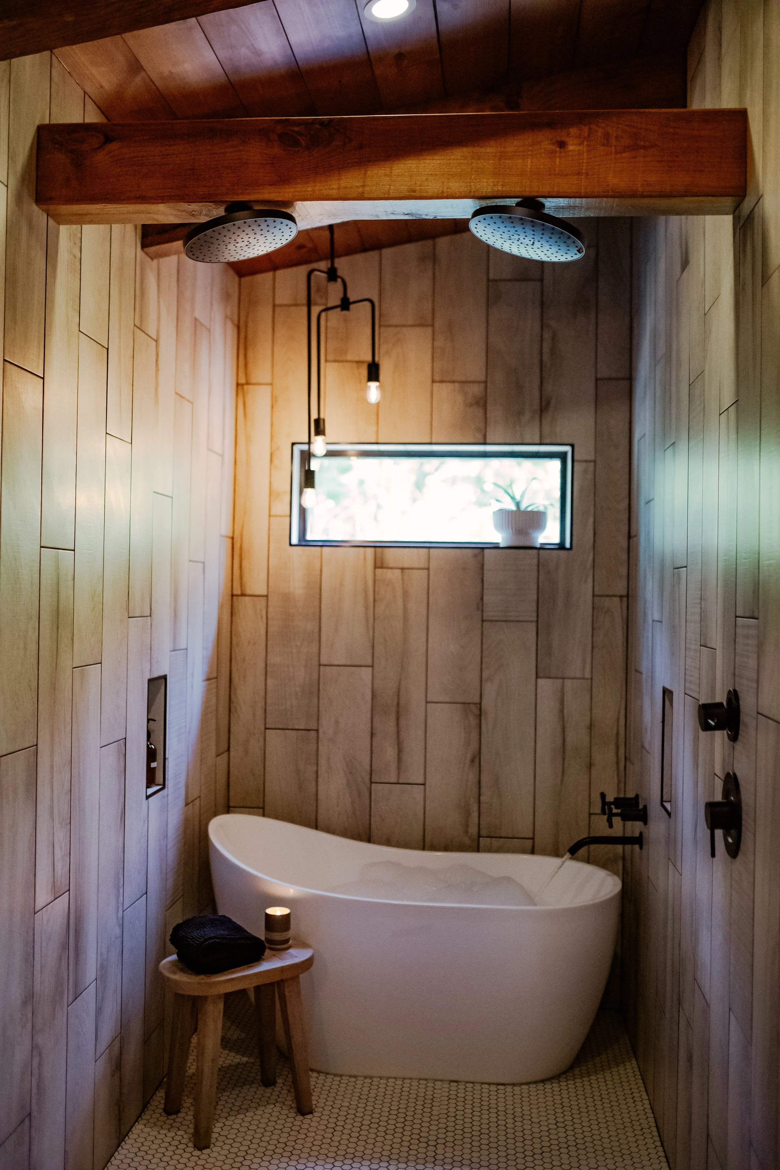 Modern bathroom with wooden wall paneling, a white freestanding bathtub, small wooden stool with a folded towel and candle, a narrow horizontal window with a potted plant, and warm lighting fixtures.