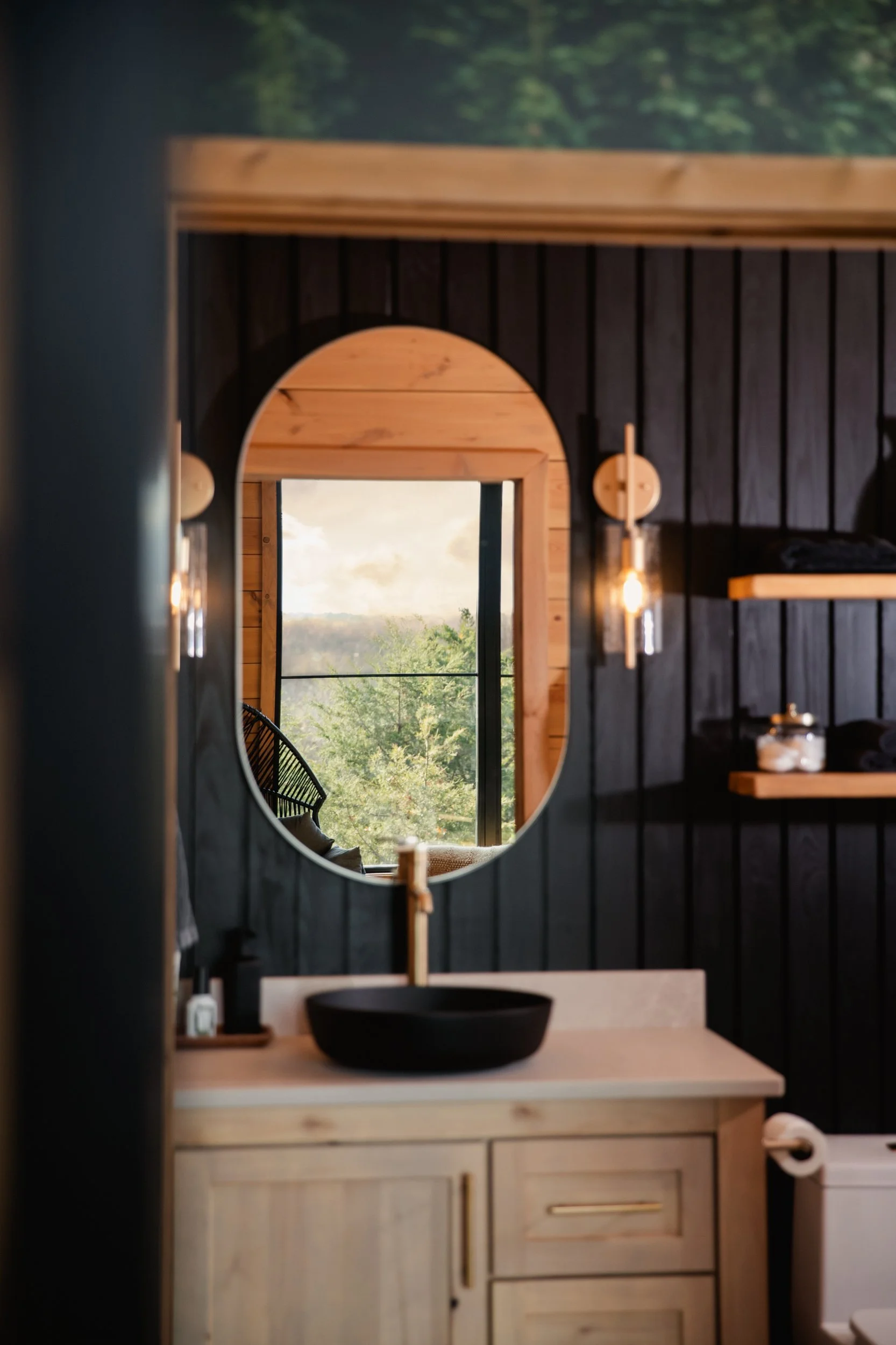Modern bathroom with a black vessel sink, wooden vanity, oval mirror, and view of trees through a large window.