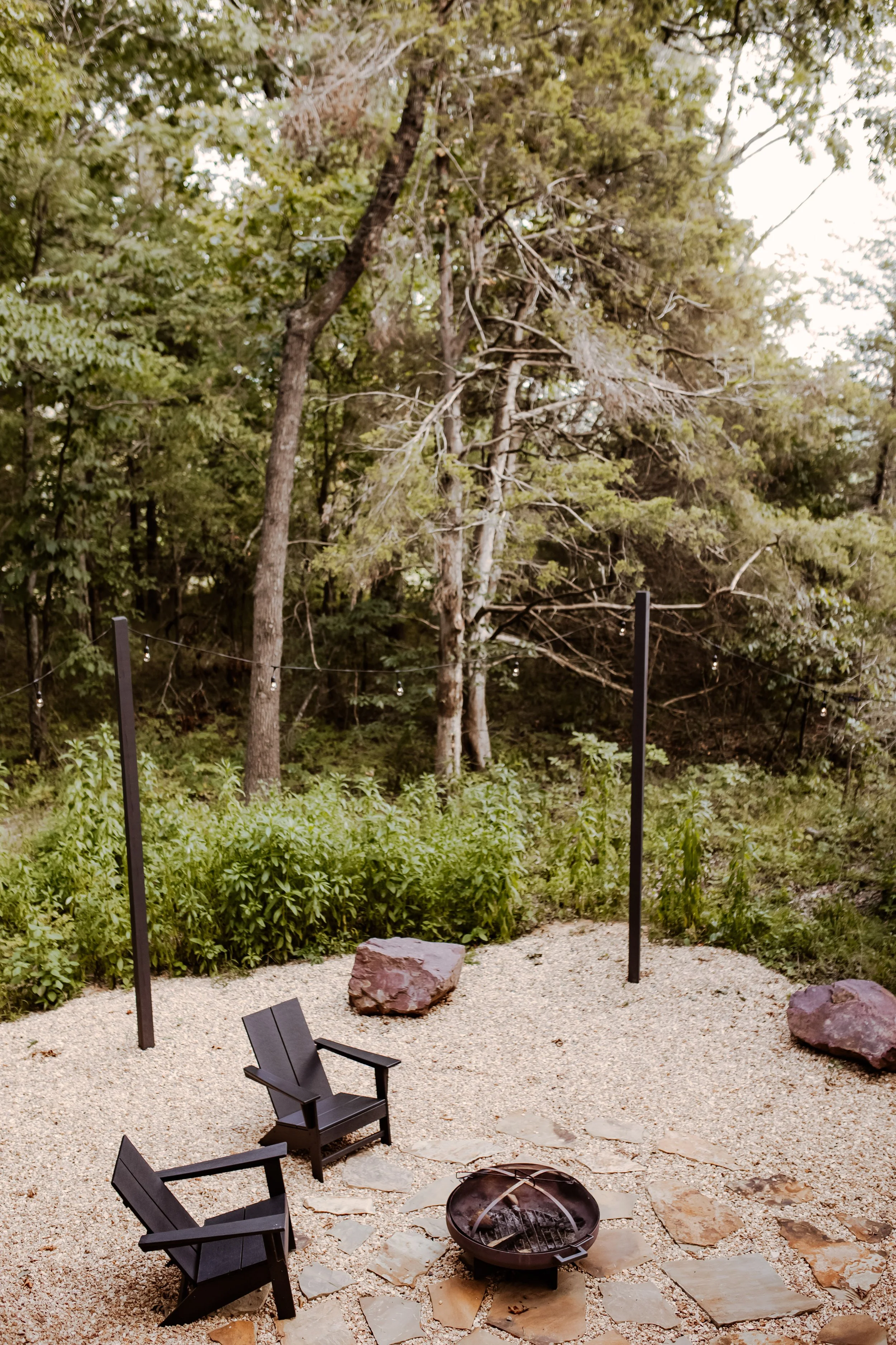 A backyard fire pit area with two black Adirondack chairs, large rocks, and a gravel ground, surrounded by green bushes and tall trees.