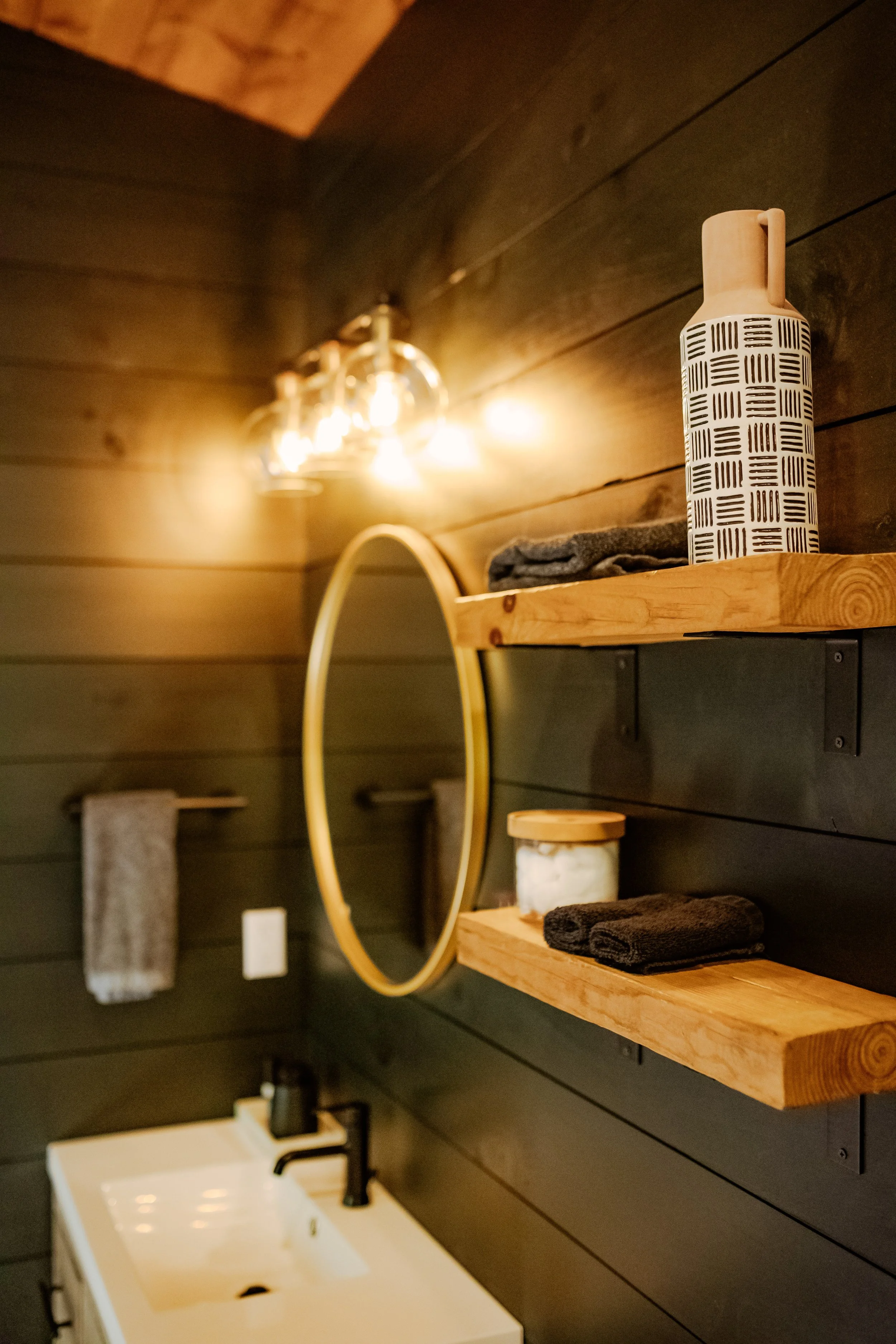 Close-up of a bathroom vanity with a white sink, black faucet, and a black soap dispenser. Behind the sink are dark wooden walls, a round mirror with a light wood frame, and two floating wooden shelves holding towels, a container of cotton balls, and