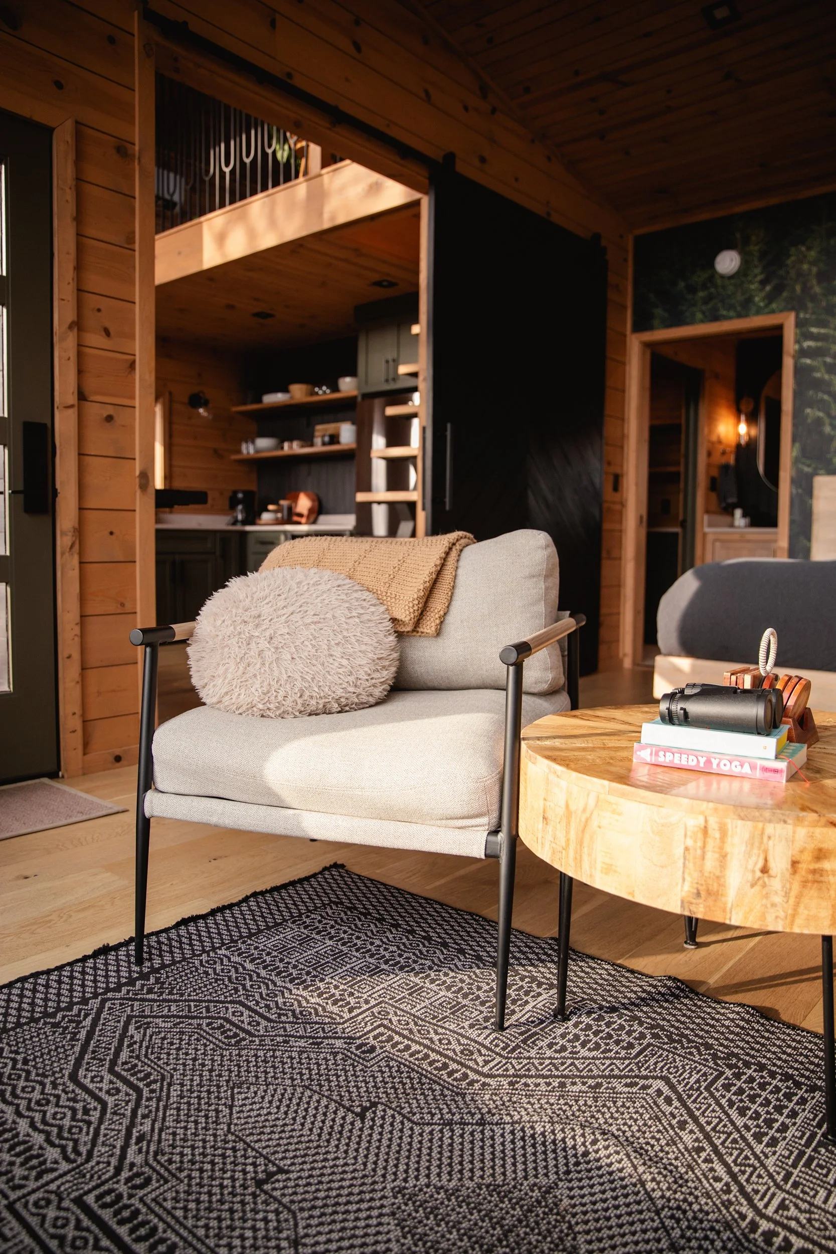 Living room with wooden walls and furniture, beige armchair with a fluffy pillow, round wooden coffee table with books and binoculars, black and white patterned rug.