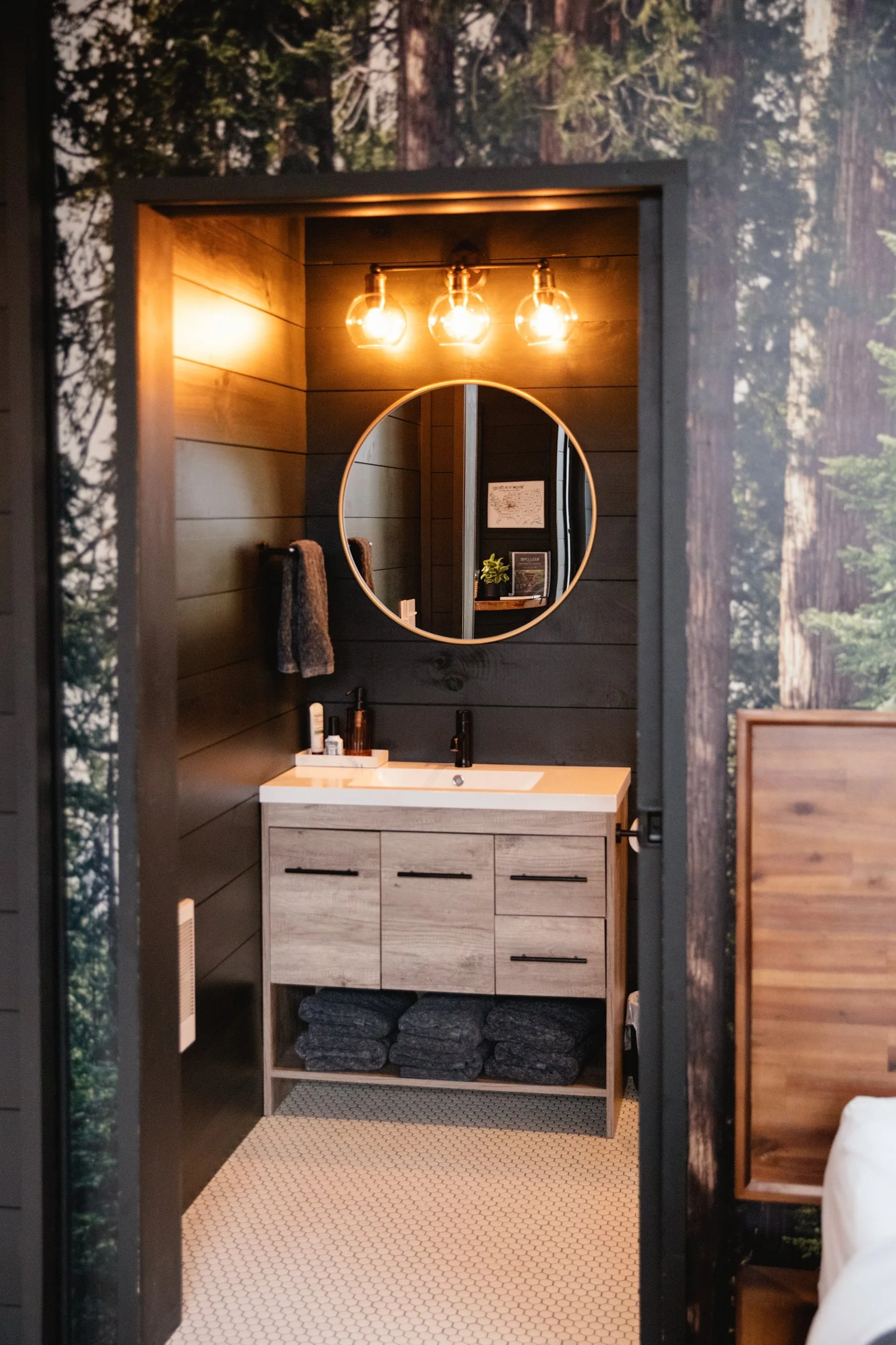 A rustic bathroom with a wood-paneled dark wall, a round mirror, three warm light bulbs, and a light-colored vanity with dark pulls. Towels, toiletries, and black stones are visible inside, with hexagonal white tiles on the floor and a nature-inspire
