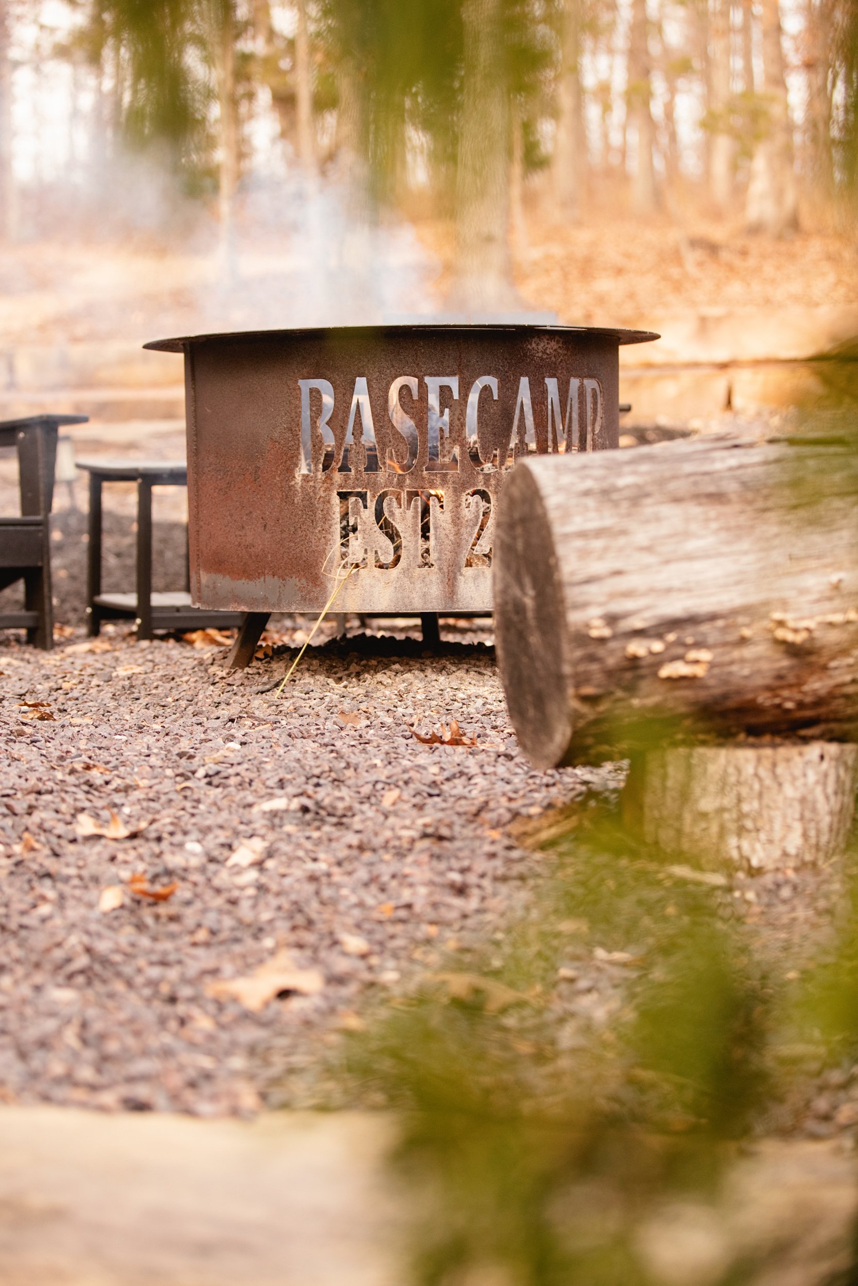 Rusty metal fire pit with cut-out text reading 'Basecamp Est 2' surrounded by trees and forest in fall, on a gravel ground with a large log in foreground.
