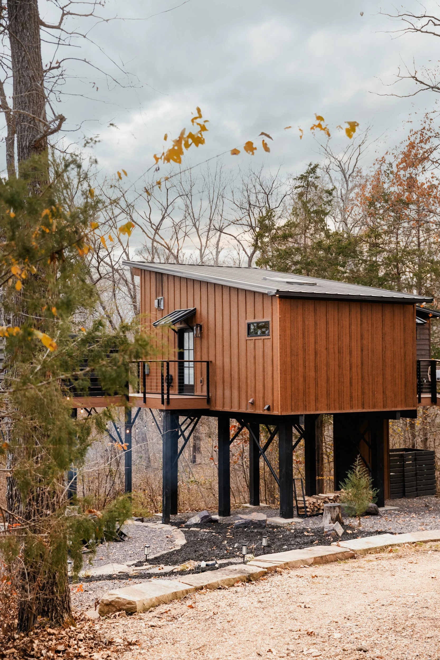 A small modern house with wooden siding, elevated on black stilts, with a metal roof and small balcony, set in an autumn wooded area.