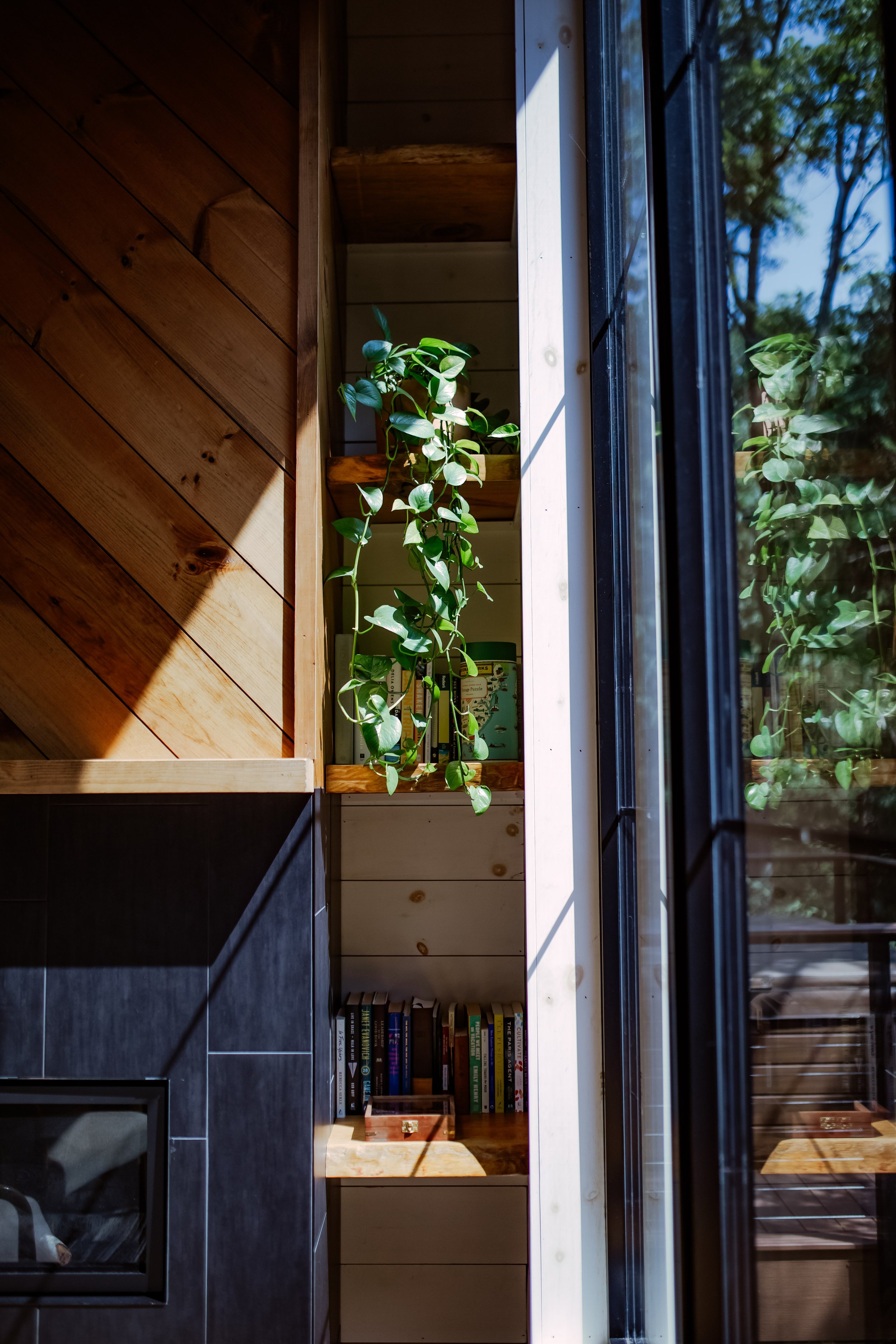 A built-in wooden bookshelf with potted plant and books, next to a large window with a view of trees.