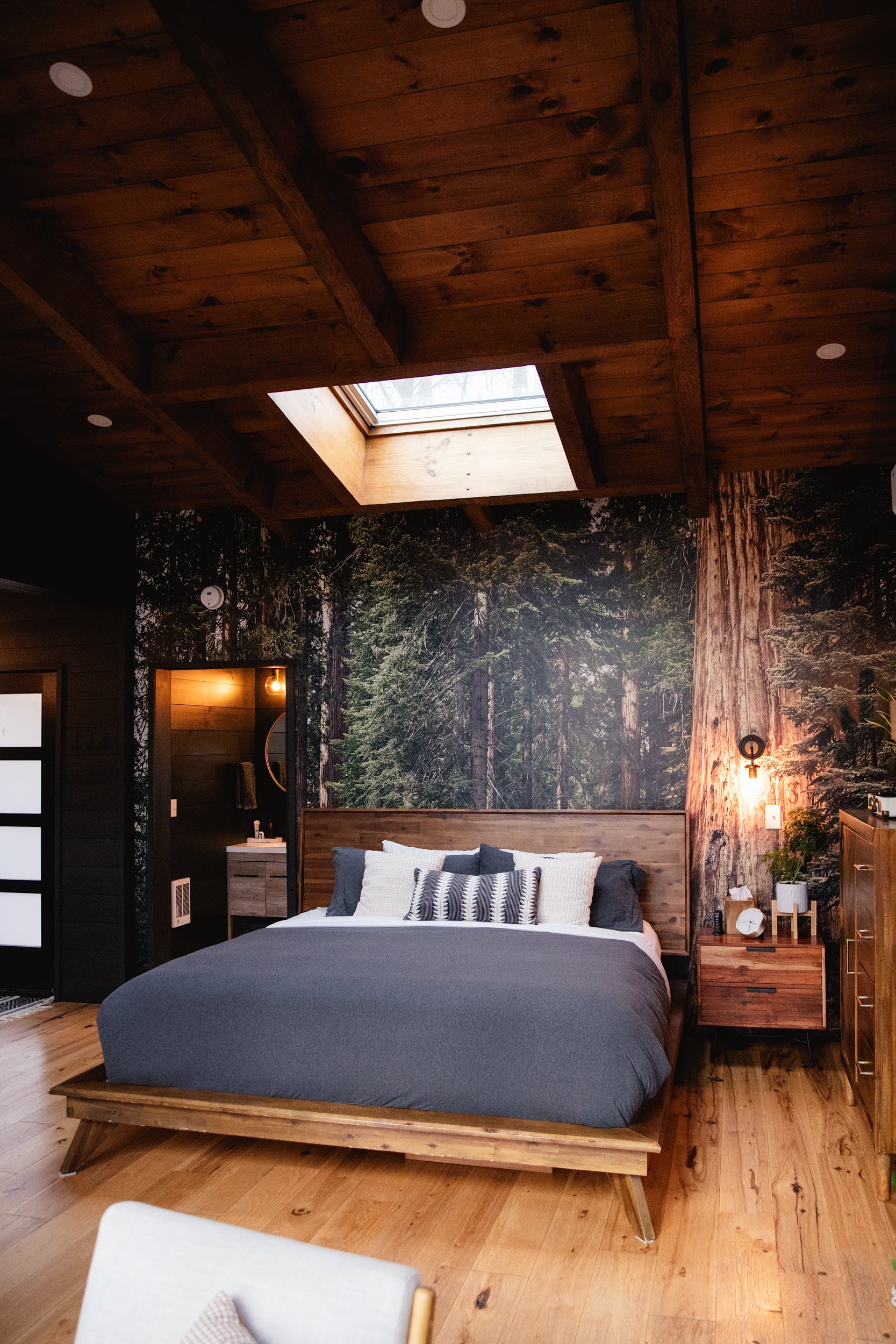 A bedroom with wooden walls and ceiling, featuring a forest mural wallpaper behind the bed. The room has a skylight in the ceiling, a wooden bed frame with dark gray bedding and white pillows, a nightstand with a plant and clock, and a small visible 