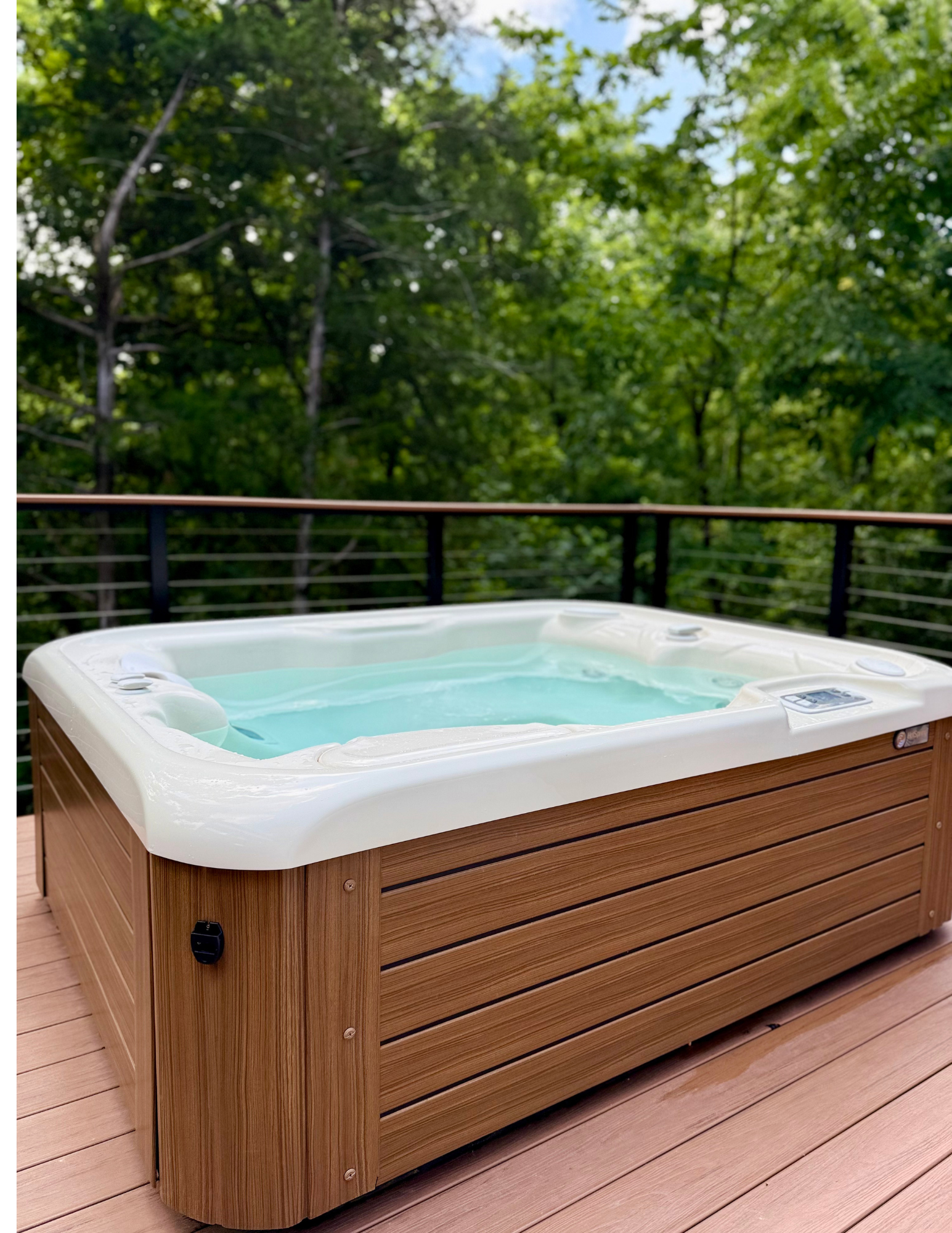 A hot tub with white interior and wooden exterior on a wooden deck, surrounded by green trees.