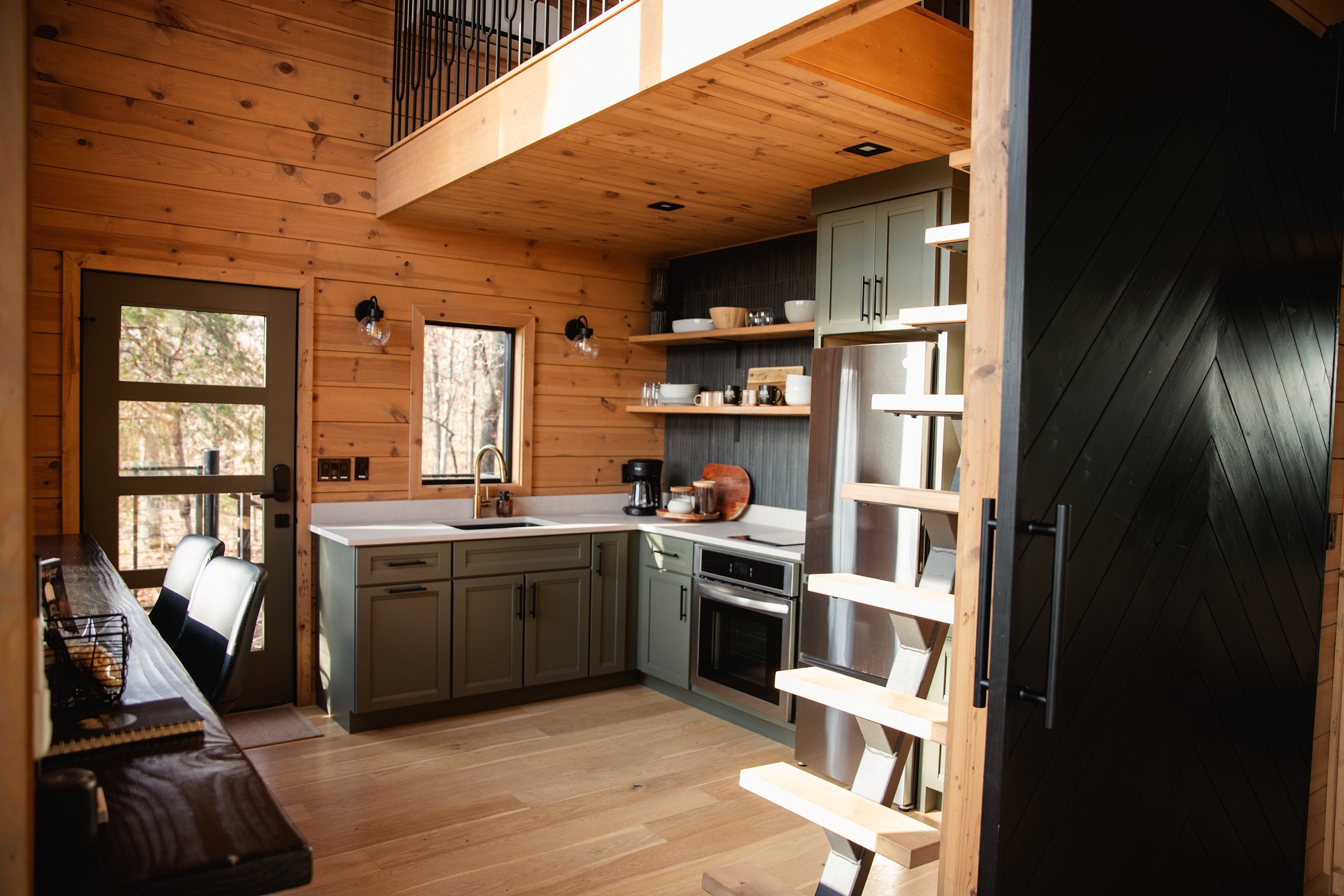 A cozy kitchen with wooden walls, green cabinets, stainless steel appliances, black accents, open shelves with dishes, and a small window above the sink.