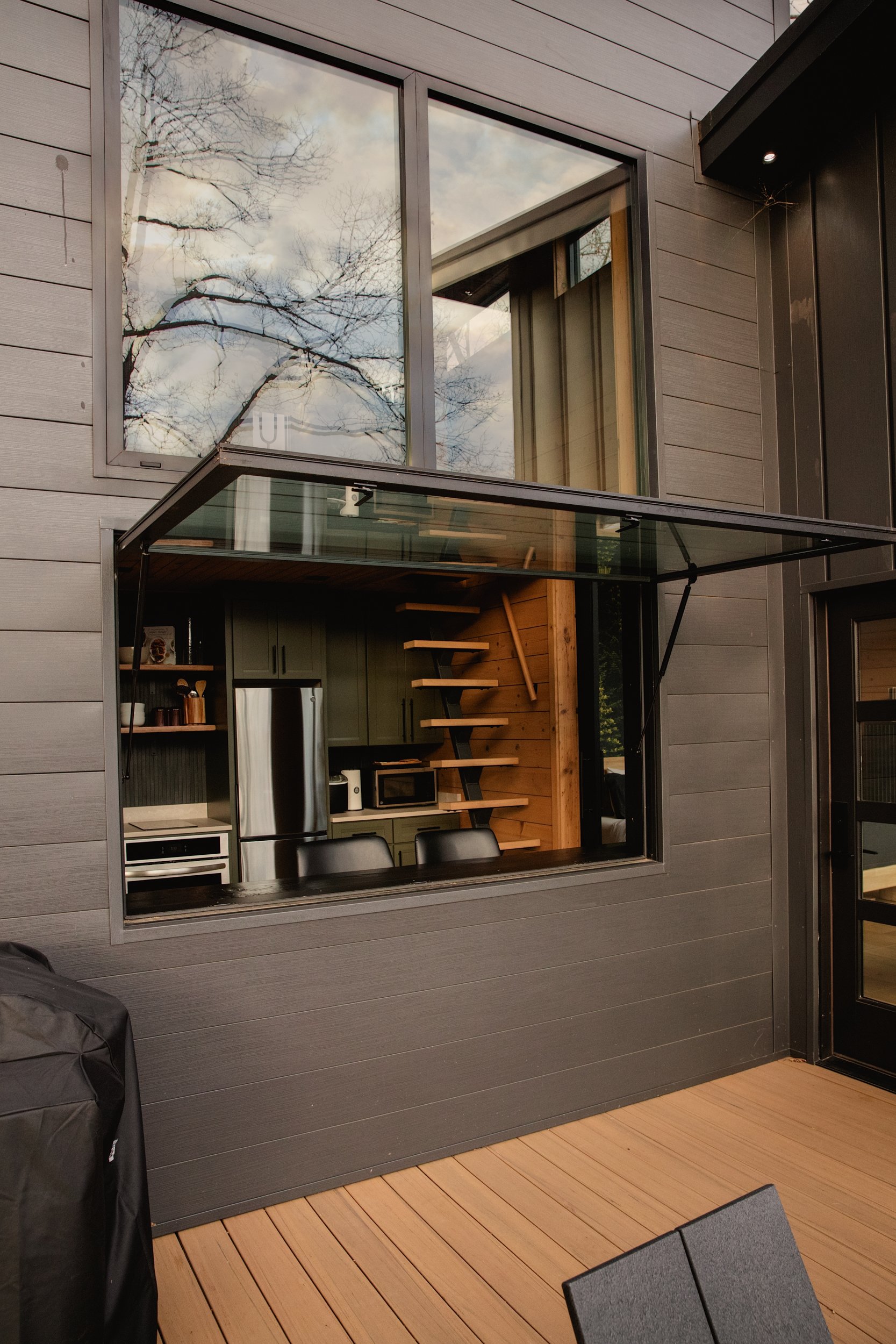 Exterior view of a modern house with a large window opening into a kitchen. The window is open, revealing kitchen appliances and a wooden staircase inside. The house has dark gray siding and a wooden deck.