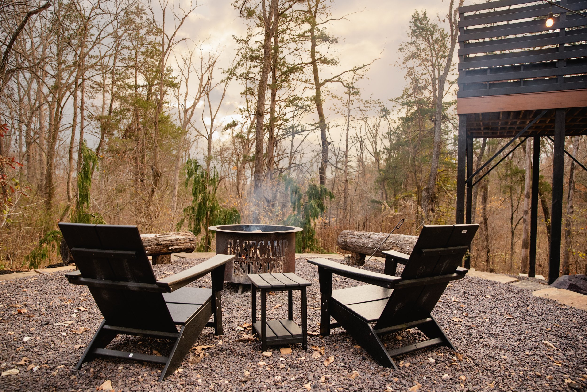 Two black outdoor chairs face a campfire ring on a gravel surface in a wooded area during autumn, with leafless trees and a cloudy sky in the background.