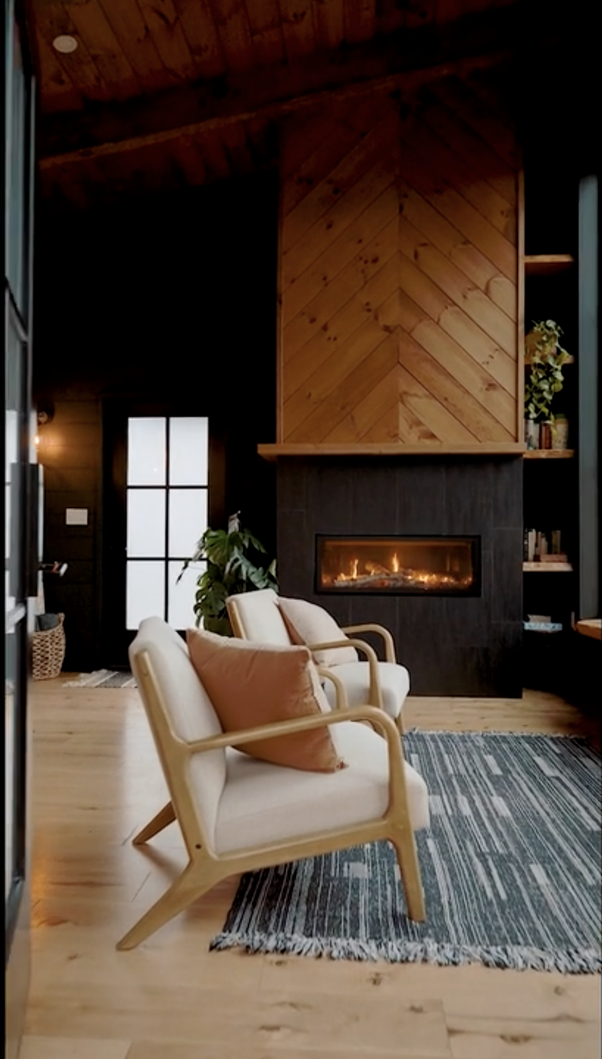 Living room with fireplace, two beige armchairs with throw pillows, a striped rug, dark wood ceiling, wooden wall on fireplace, bookshelves, potted plant, and door with frosted glass panes.
