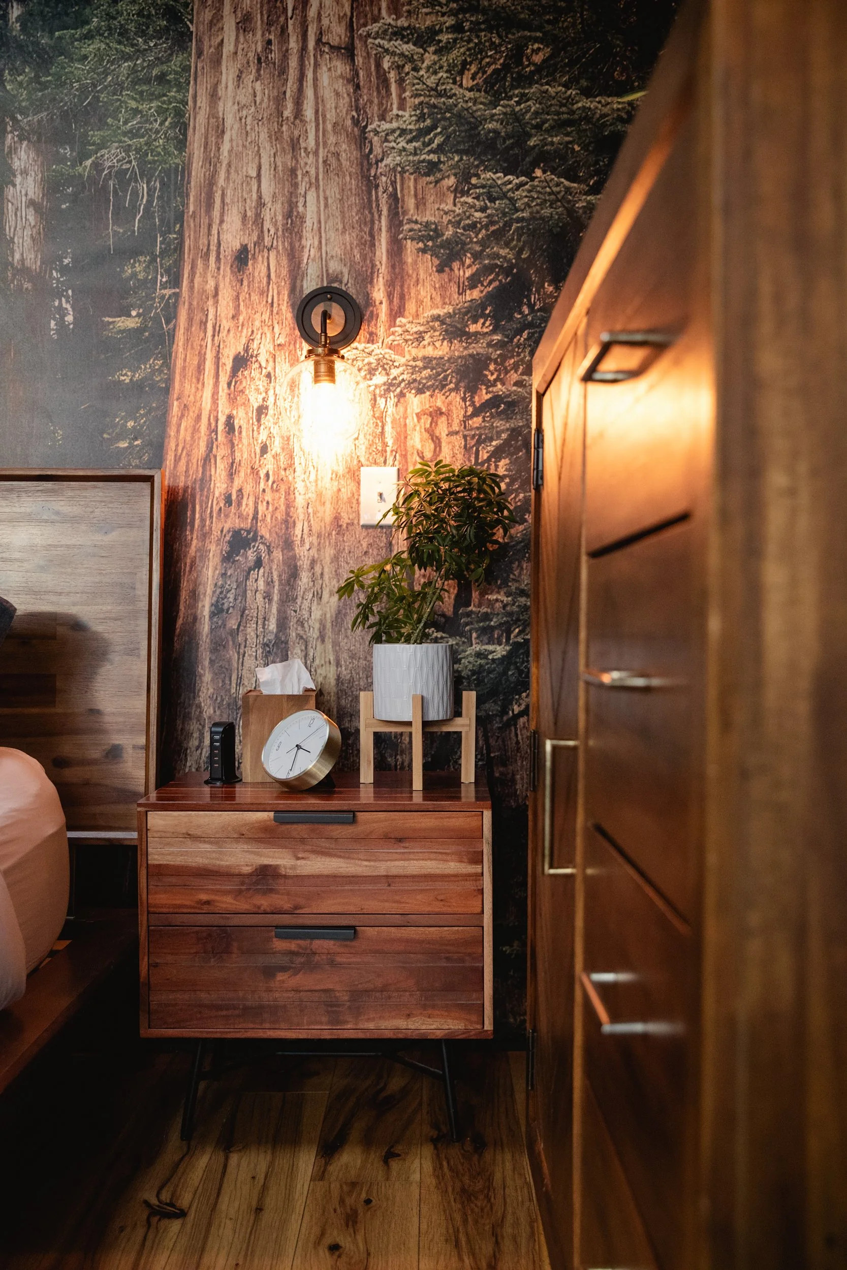 A side view of a wooden bedroom nightstand with a black and gold clock, a small black alarm clock, and a potted plant on top, against a wall with a forest mural, a wall-mounted light bulb, and a power outlet.