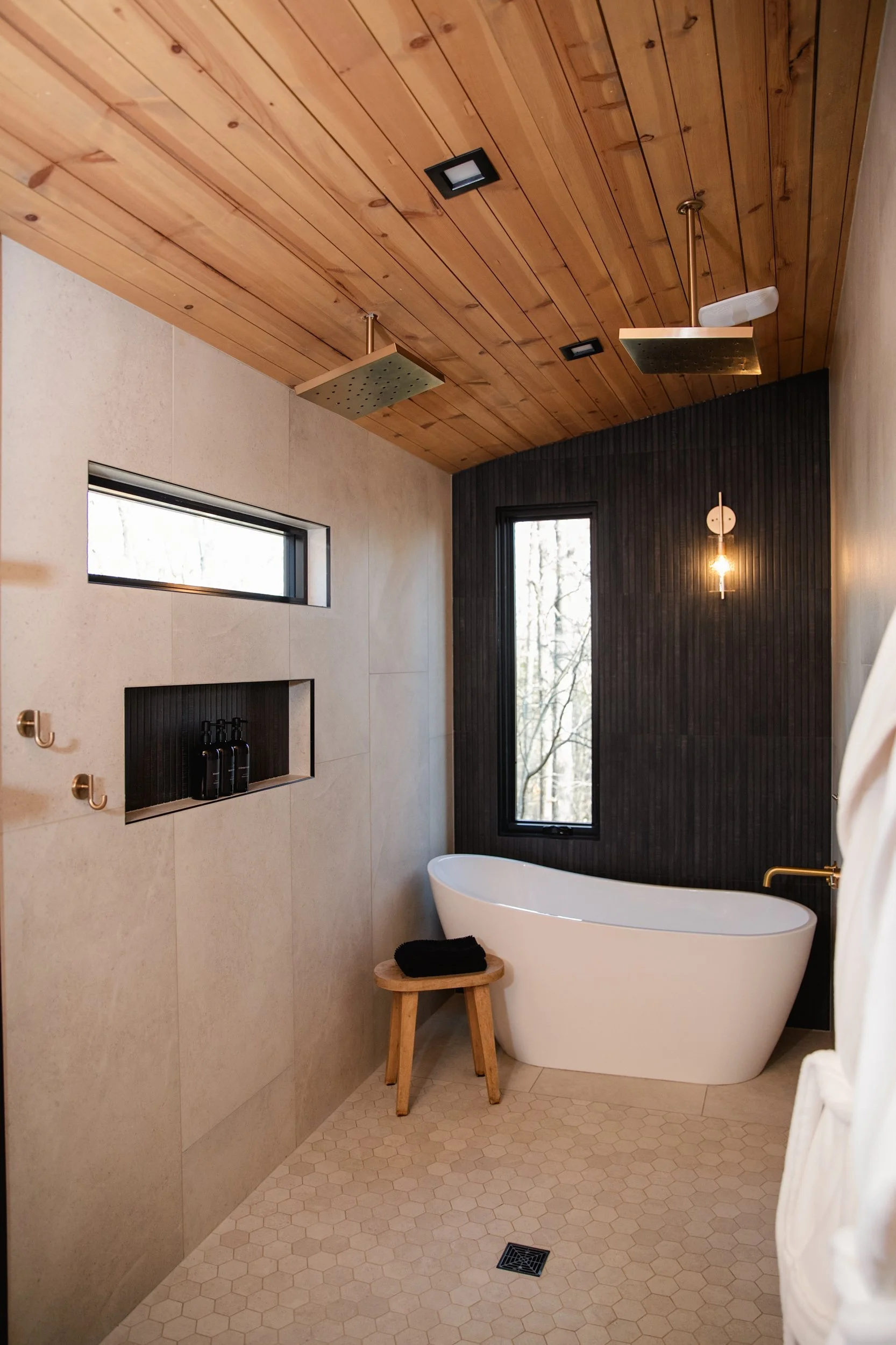 Modern bathroom with a white bathtub, black accent wall, wooden ceiling, and a large window with a view of trees.
