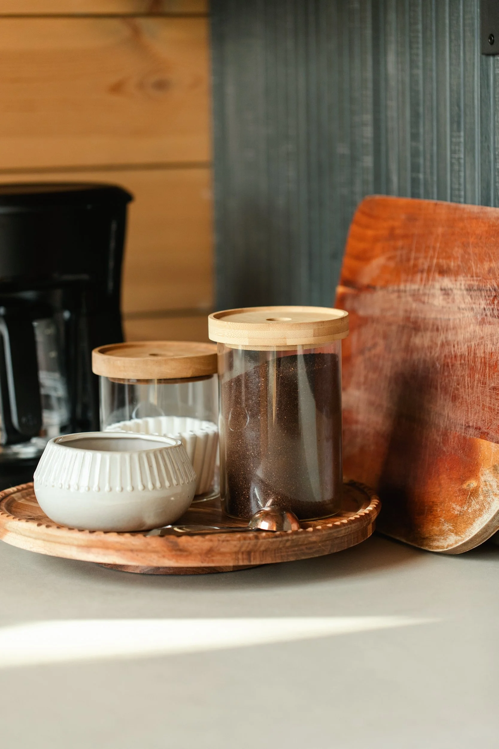 Coffee and tea jars on a wooden tray with a small jug and a spoon, positioned on a countertop.