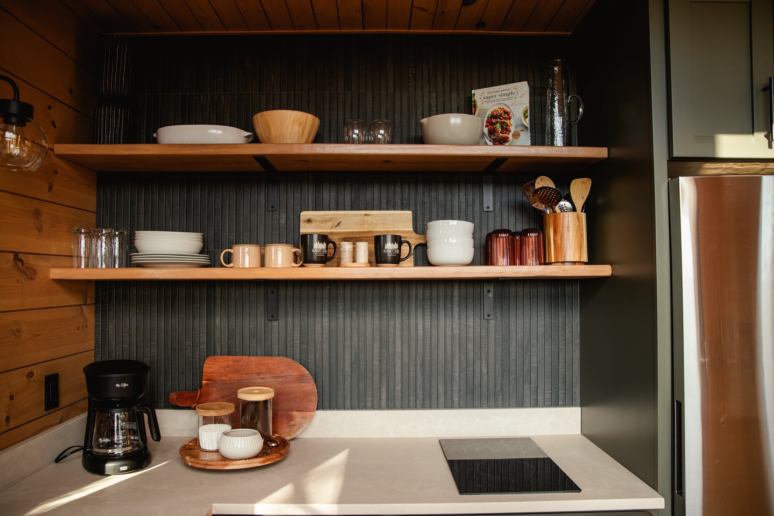 Open kitchen shelf with bowls, glasses, mugs, and kitchen utensils. Coffee maker, cutting board, and small dishes on counter.
