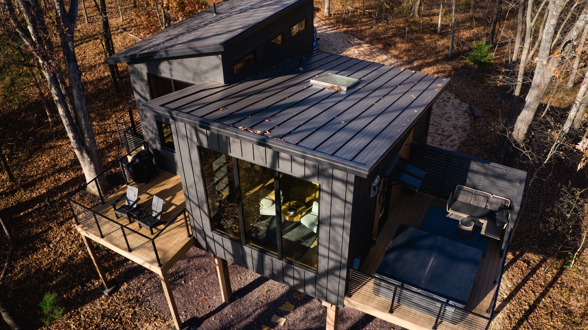 Aerial view of a modern, small house with a black metal roof, large windows, and outdoor decks in a wooded area with deciduous trees.