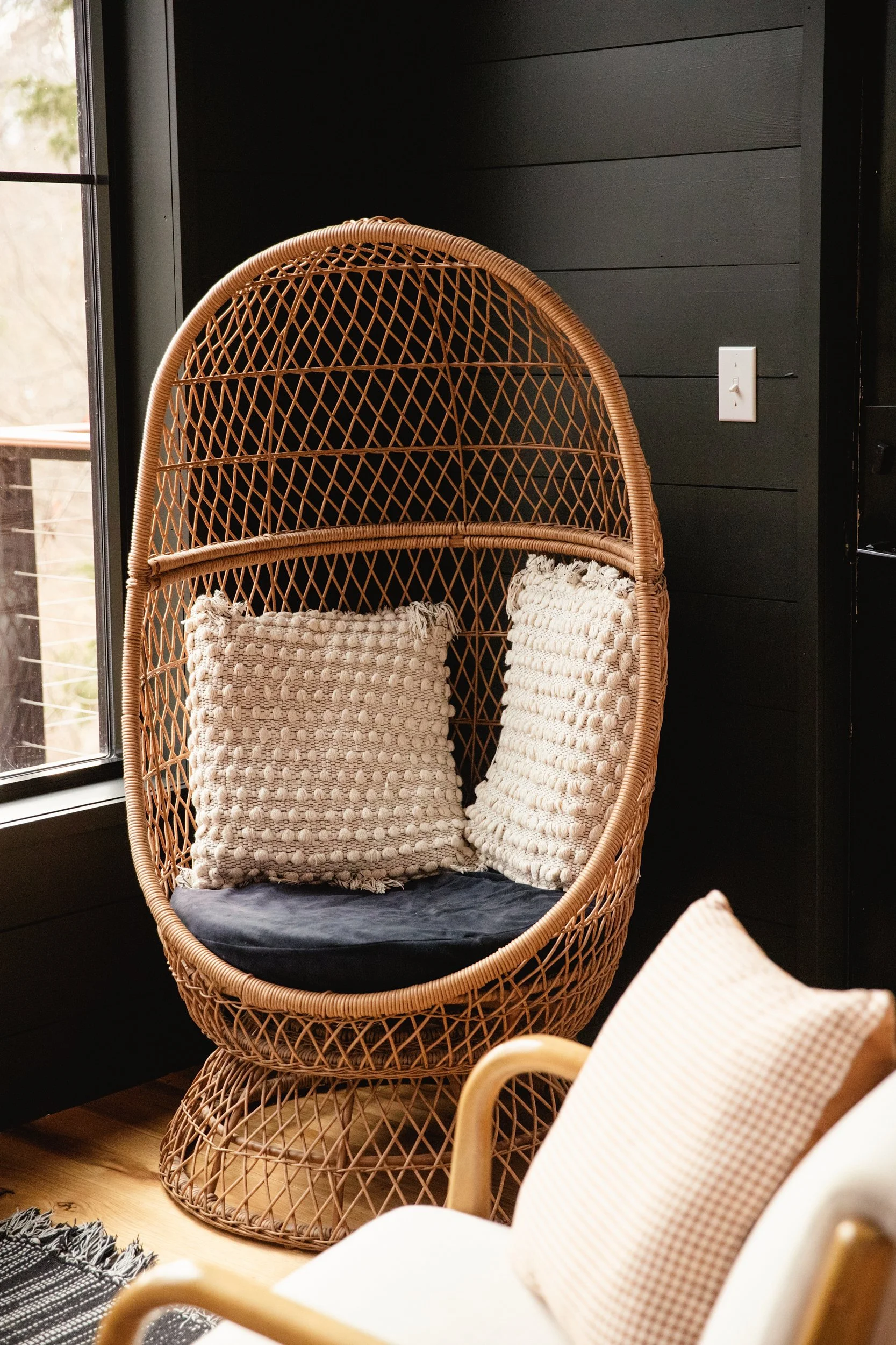 Wicker egg-shaped chair with two textured throw pillows, black seat cushion, near a window and black wall.
