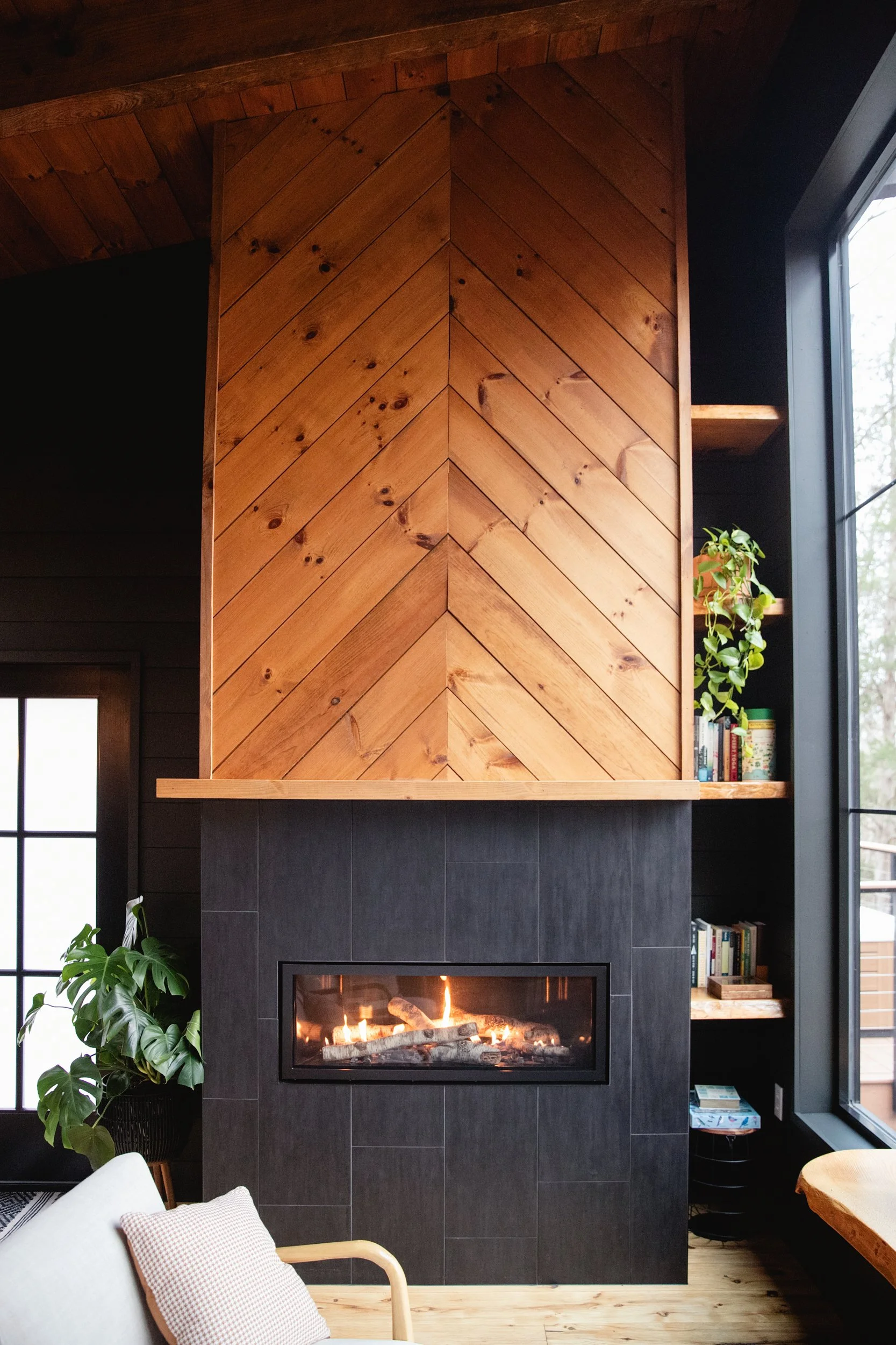 Indoor room featuring a modern fireplace with black tiles and a wood-paneled chimney, surrounded by built-in shelves with books and a potted plant, next to a large window.