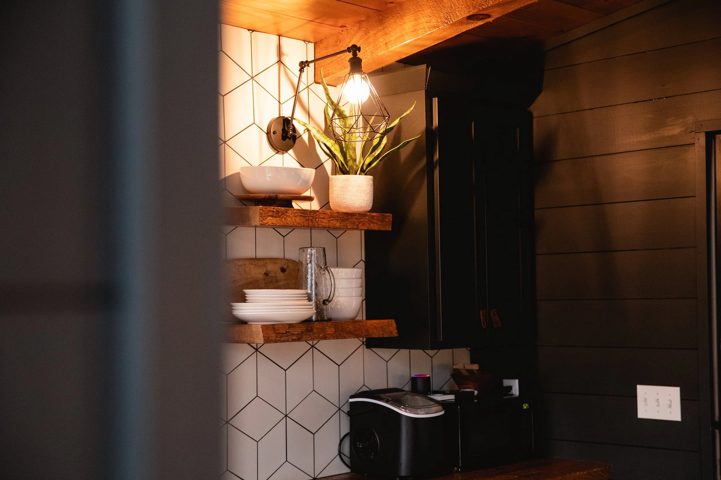 Kitchen with wooden shelves holding white dishes and a potted plant, black cabinets, and hexagonal white tiles.