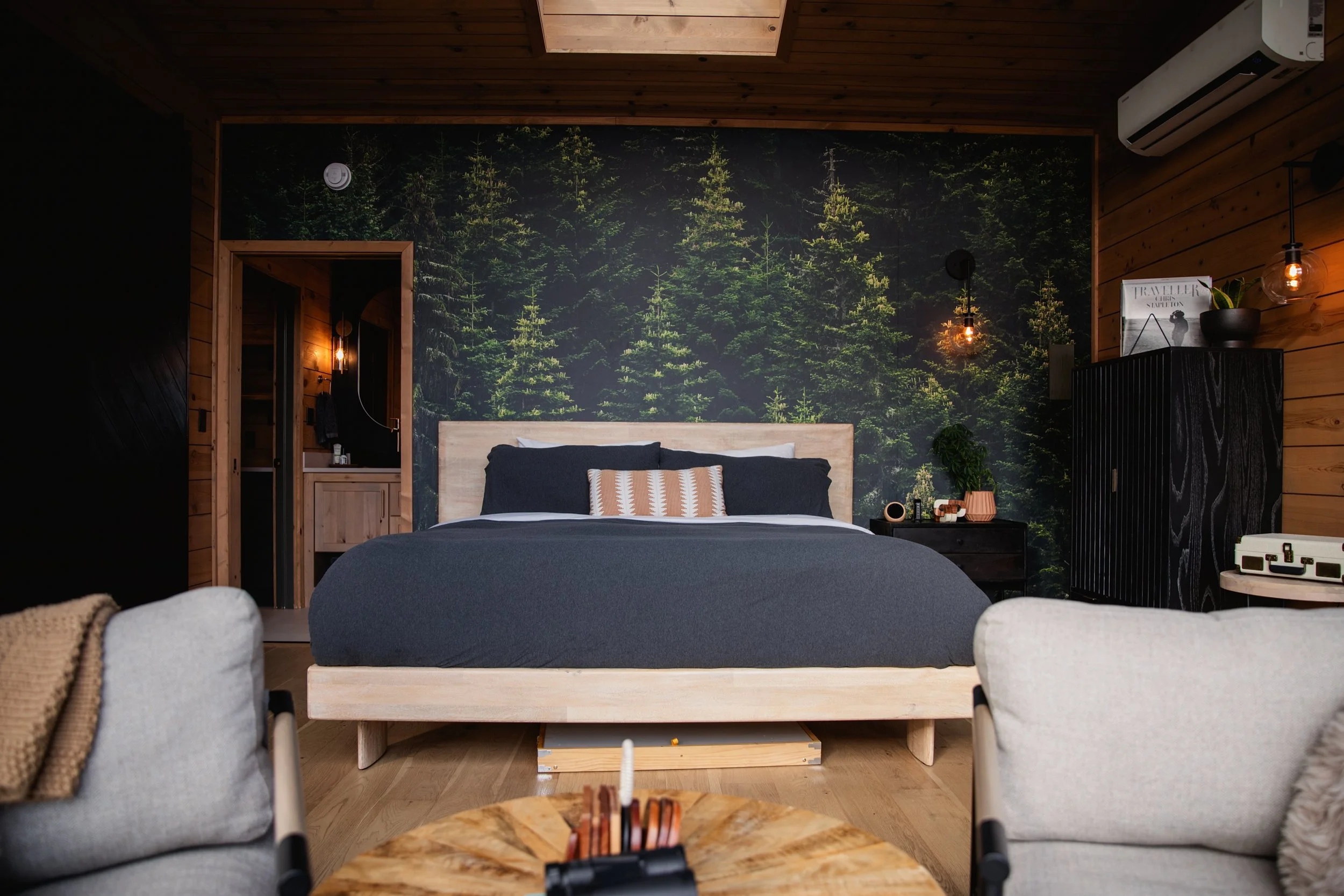 Interior of a cozy bedroom with a forest-themed wallpaper, wooden ceiling, and modern furniture. A bed with dark gray bedding and a striped pillow is centered, with bedside table and decorative items. Two armchairs are in the foreground, and a small 