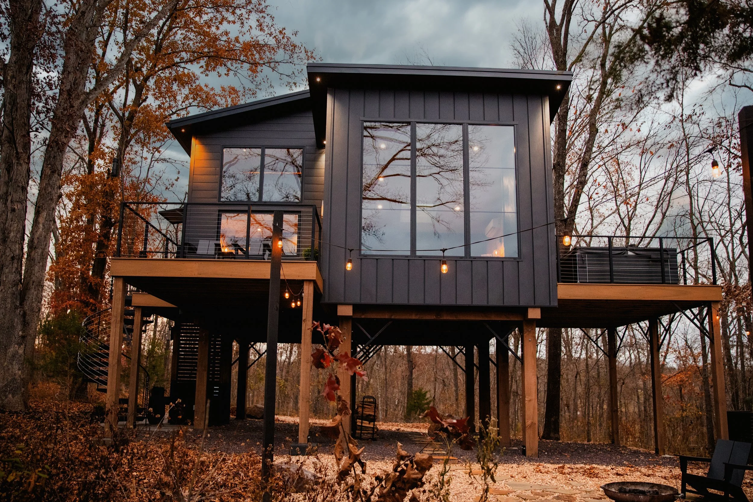 Modern two-story house on stilts with large windows, black siding, string lights, and outdoor seating in a wooded area during dusk.