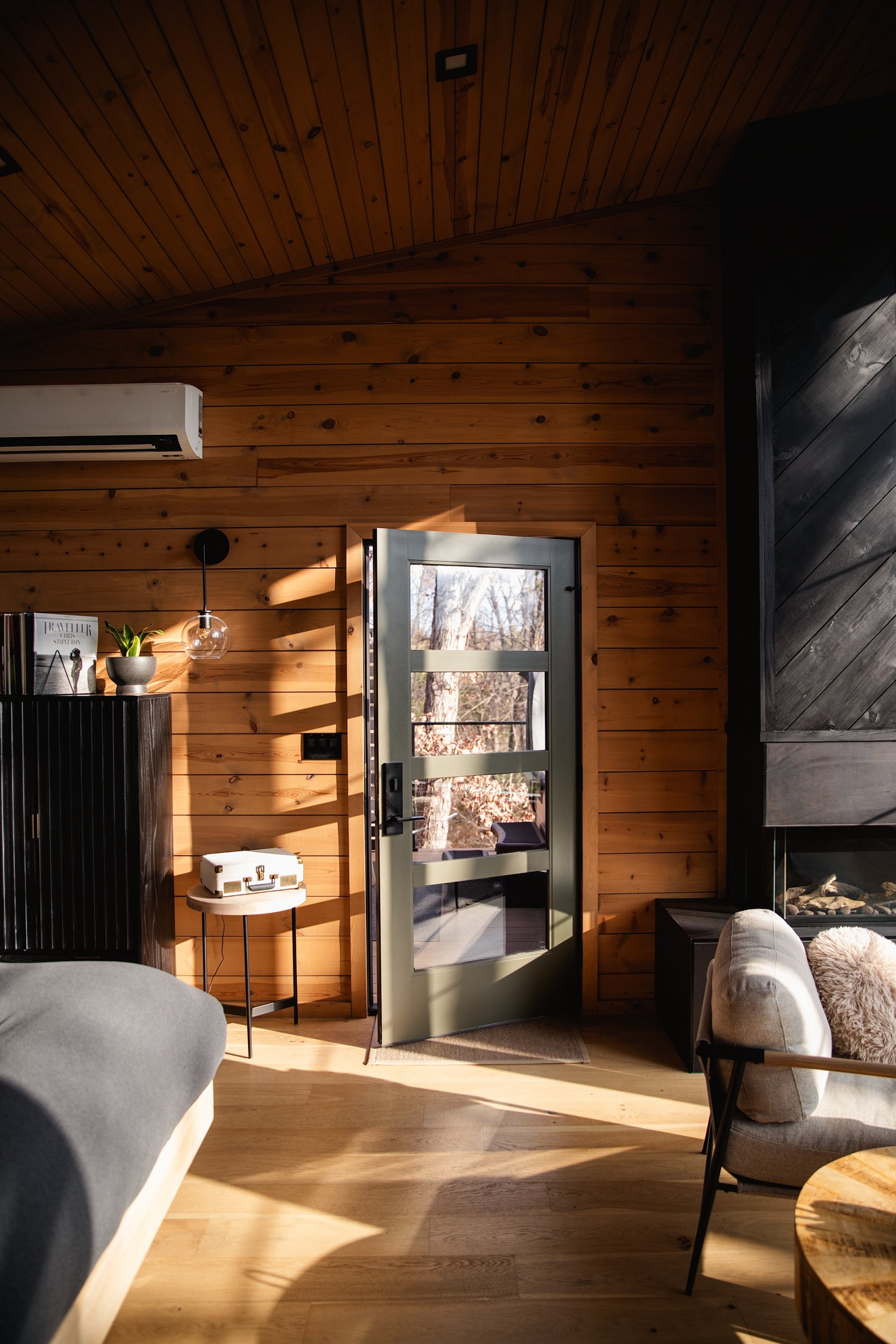 Living room with wood-paneled walls and ceiling, a glass door leading outside, a sofa with light upholstery, a chair with a fur pillow, a black side table, a small white suitcase, and a bookshelf with decorative items, sunlight streaming in.