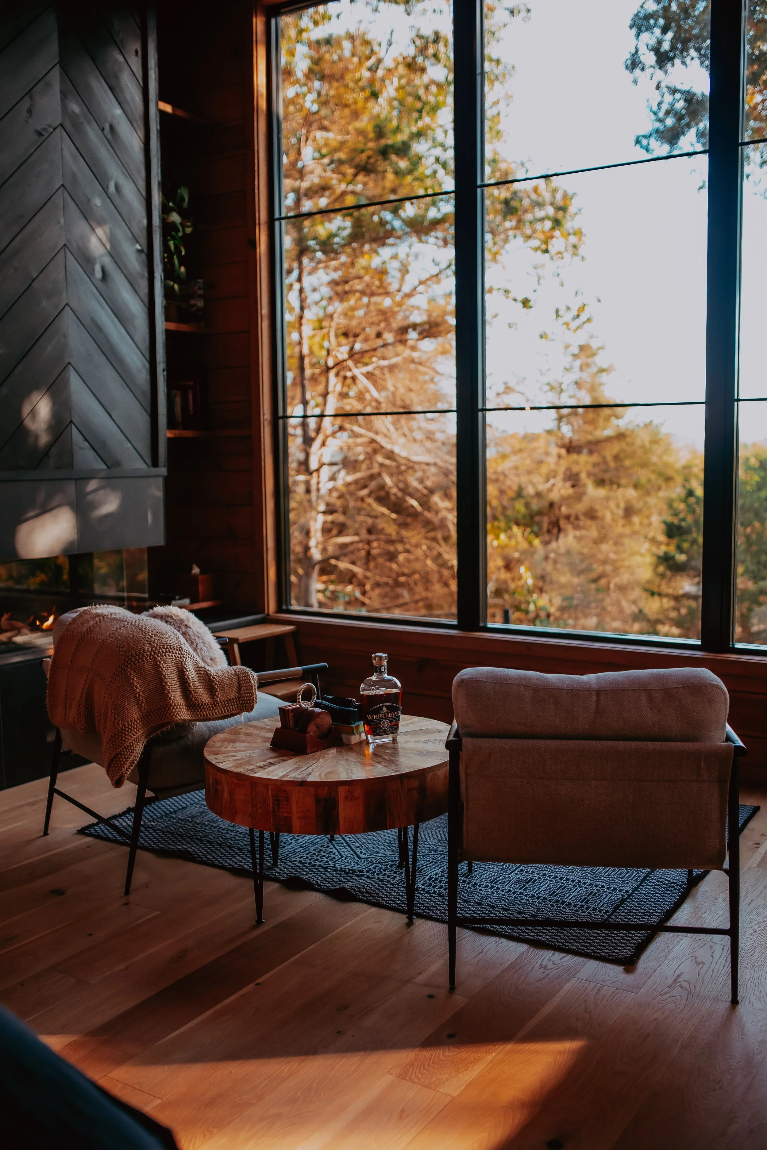 Cozy living room with two chairs and a round wooden coffee table, large window showing trees with autumn foliage, a blanket draped over one chair, and a bottle on the table.