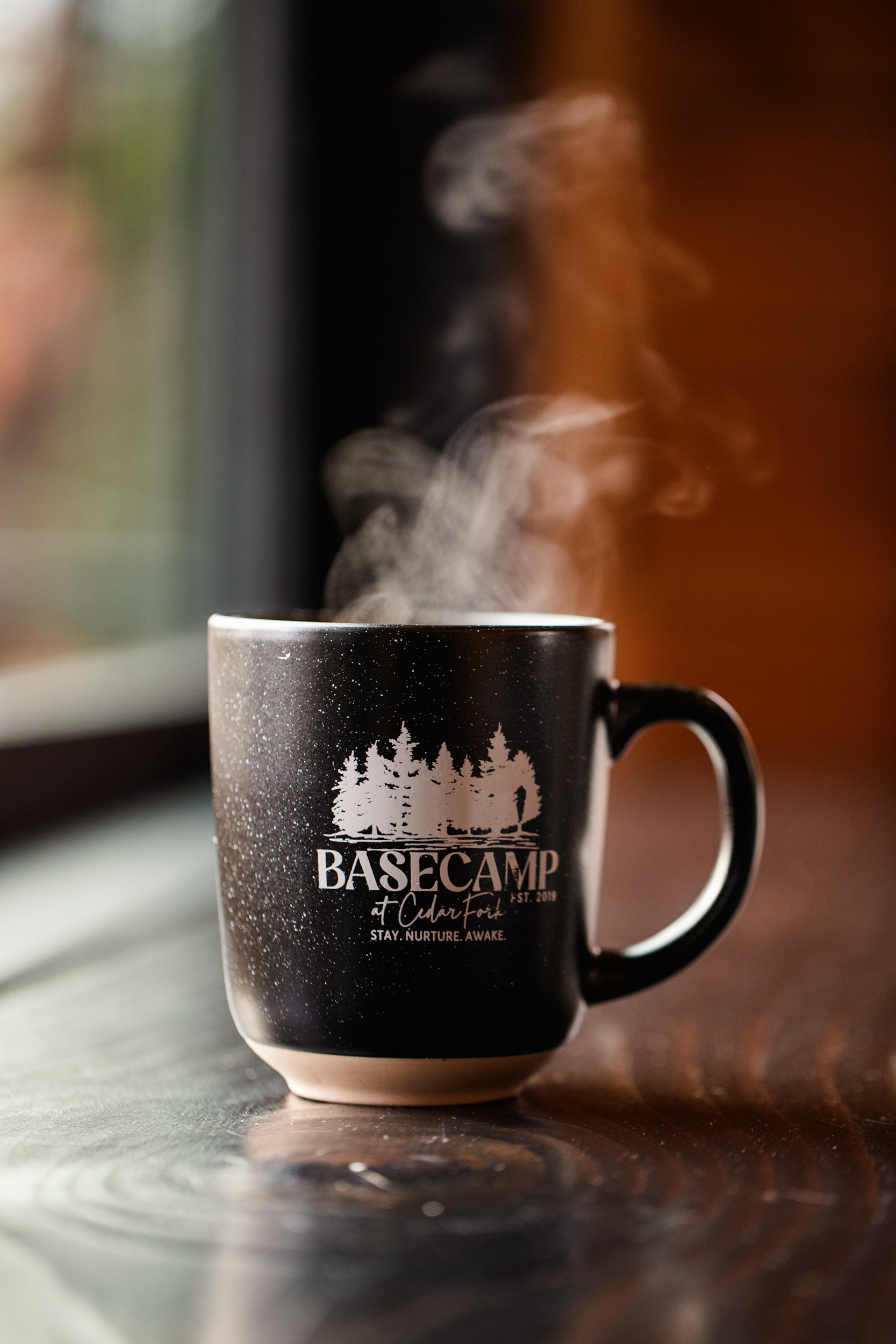 Black ceramic mug with white mountain and forest design, labeled 'BASECAMP at Charter Oak, EST. 2019, STAY. NURTURE. AWAKE.', with steam rising from hot beverage inside, placed on a wooden surface near a window.