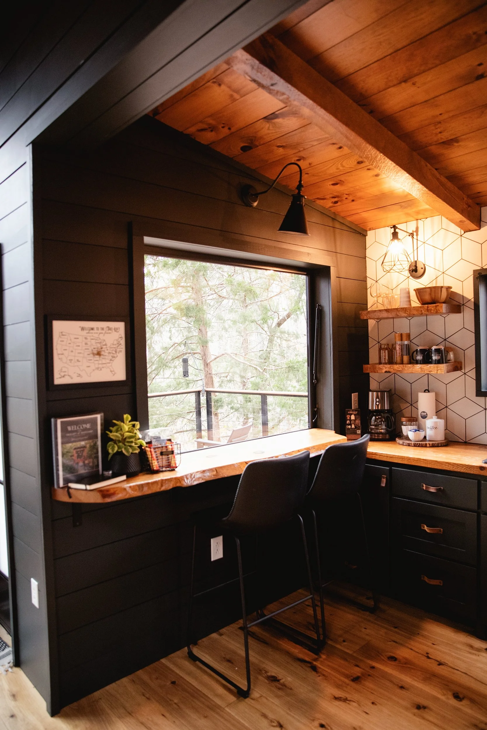 A cozy kitchen corner with black walls and wooden accents, featuring a large window overlooking trees, a wooden bar counter with two black chairs, open shelves with kitchen items, and warm lighting.