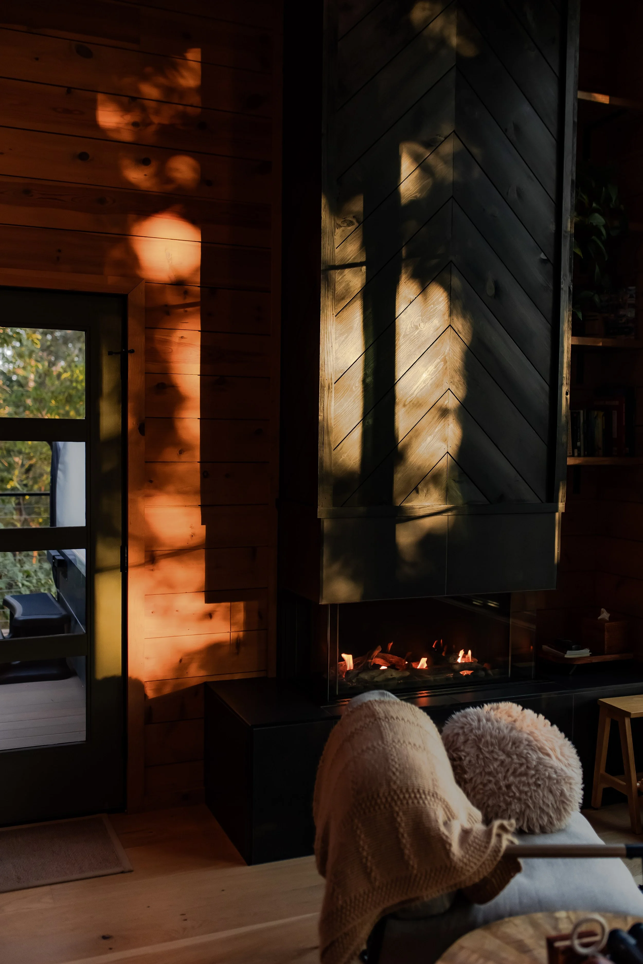Cozy living room interior with a lit fireplace, wooden walls, a door with glass panes leading outside, and the warm glow of sunlight casting shadows on the wall. A chair with a fluffy pillow and blanket is in the foreground.