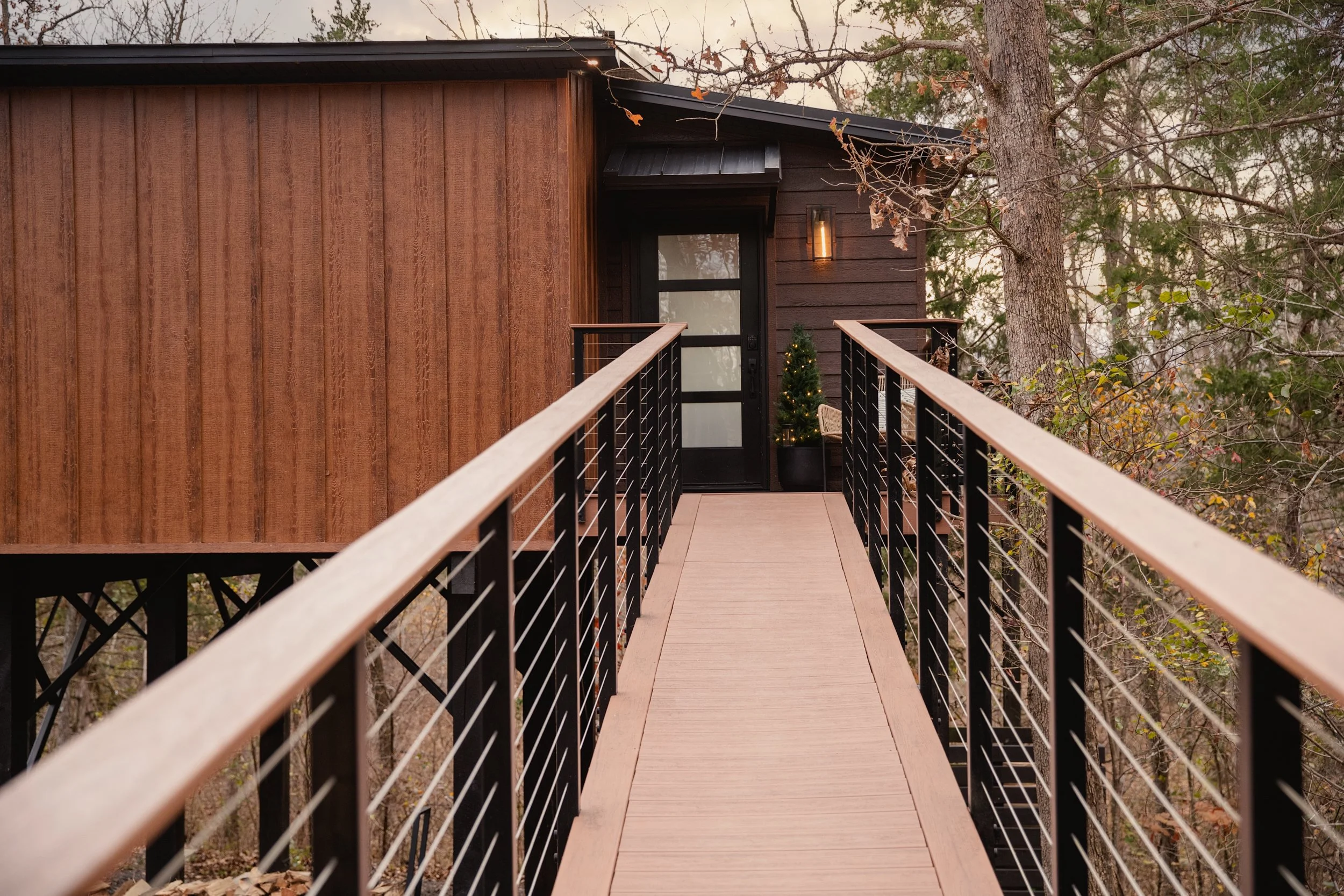 Handicap accessible walkway leading to a treehouse with a black door, surrounded by trees with some autumn leaves, decorated with a small Christmas tree outside.