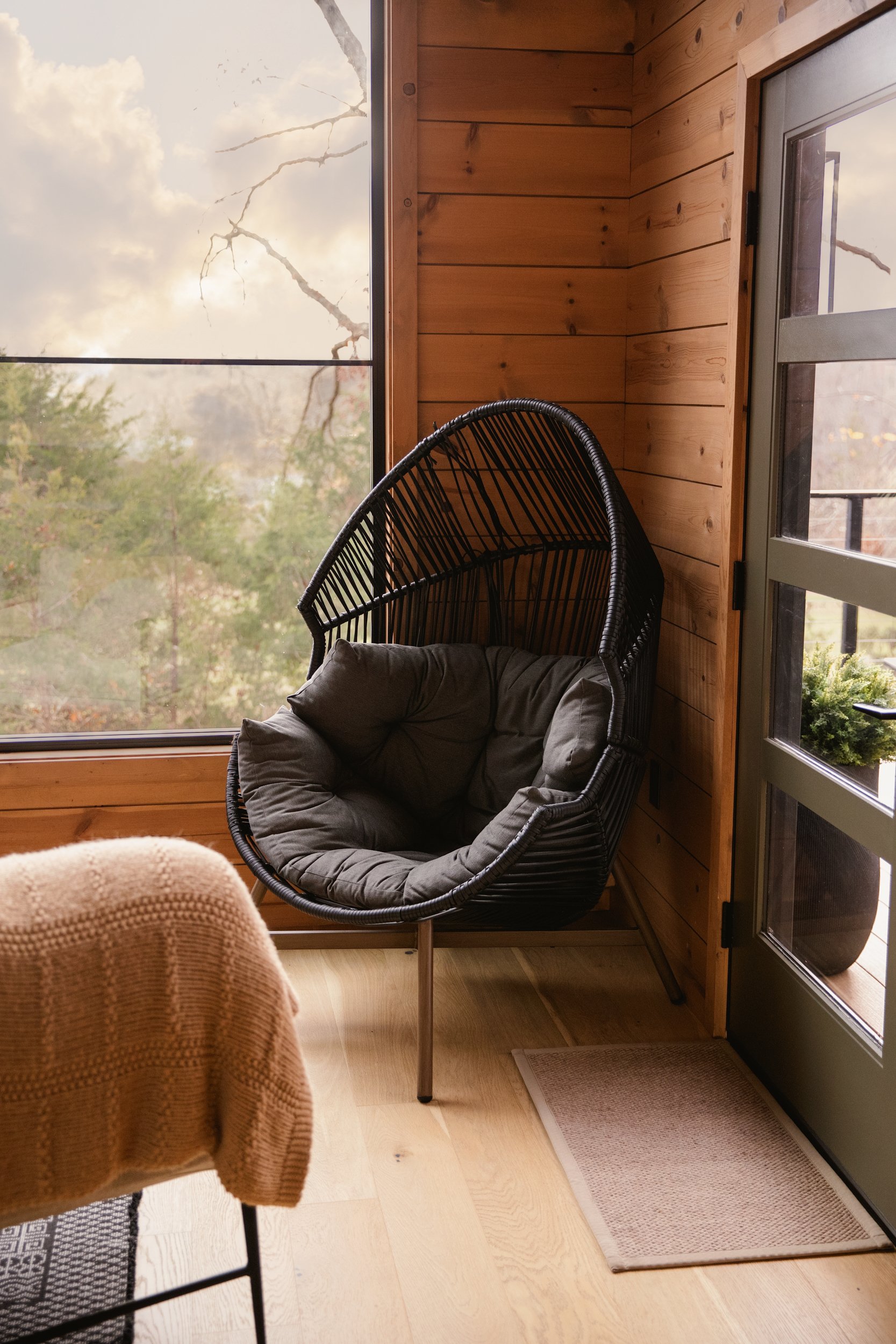 A cozy indoor corner with a black hanging egg chair with a dark cushion, positioned near a large window showing an outdoor view of trees and cloudy sky, with wooden walls and a small gray mat on the floor.