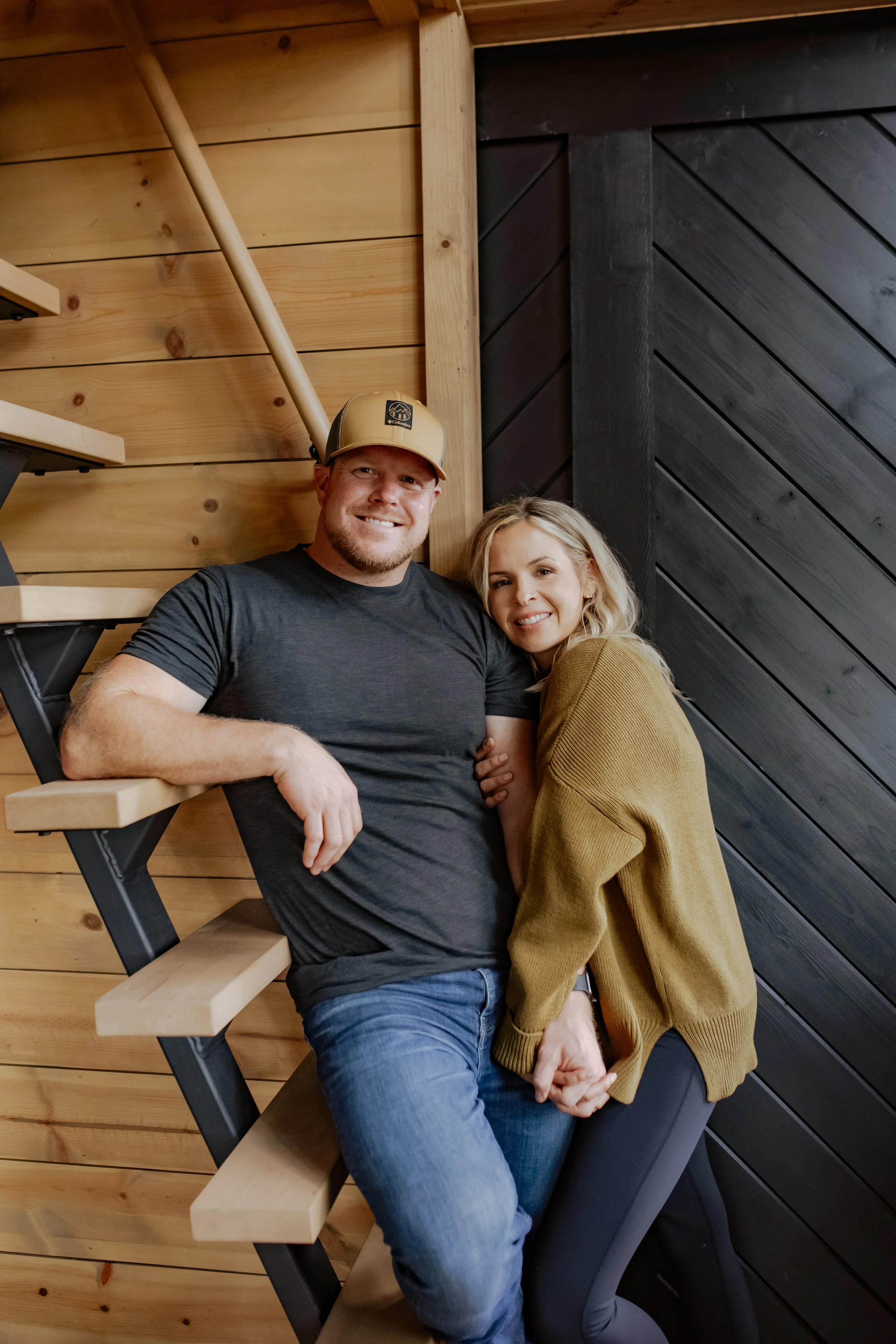 A couple sitting on stairs inside a wooden house, smiling at the camera, holding hands.