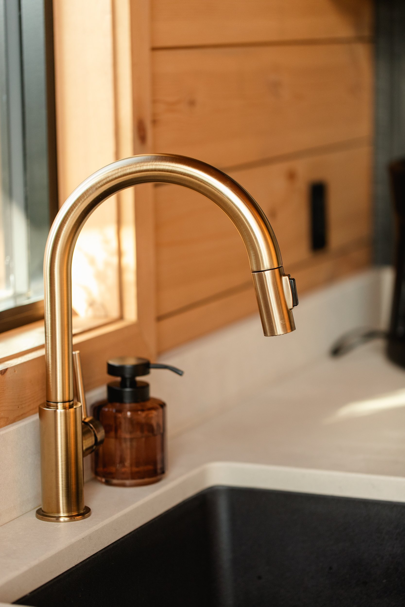 Close-up of a gold kitchen faucet over a black sink, with a brown soap dispenser on the counter and wooden cabinets in the background.