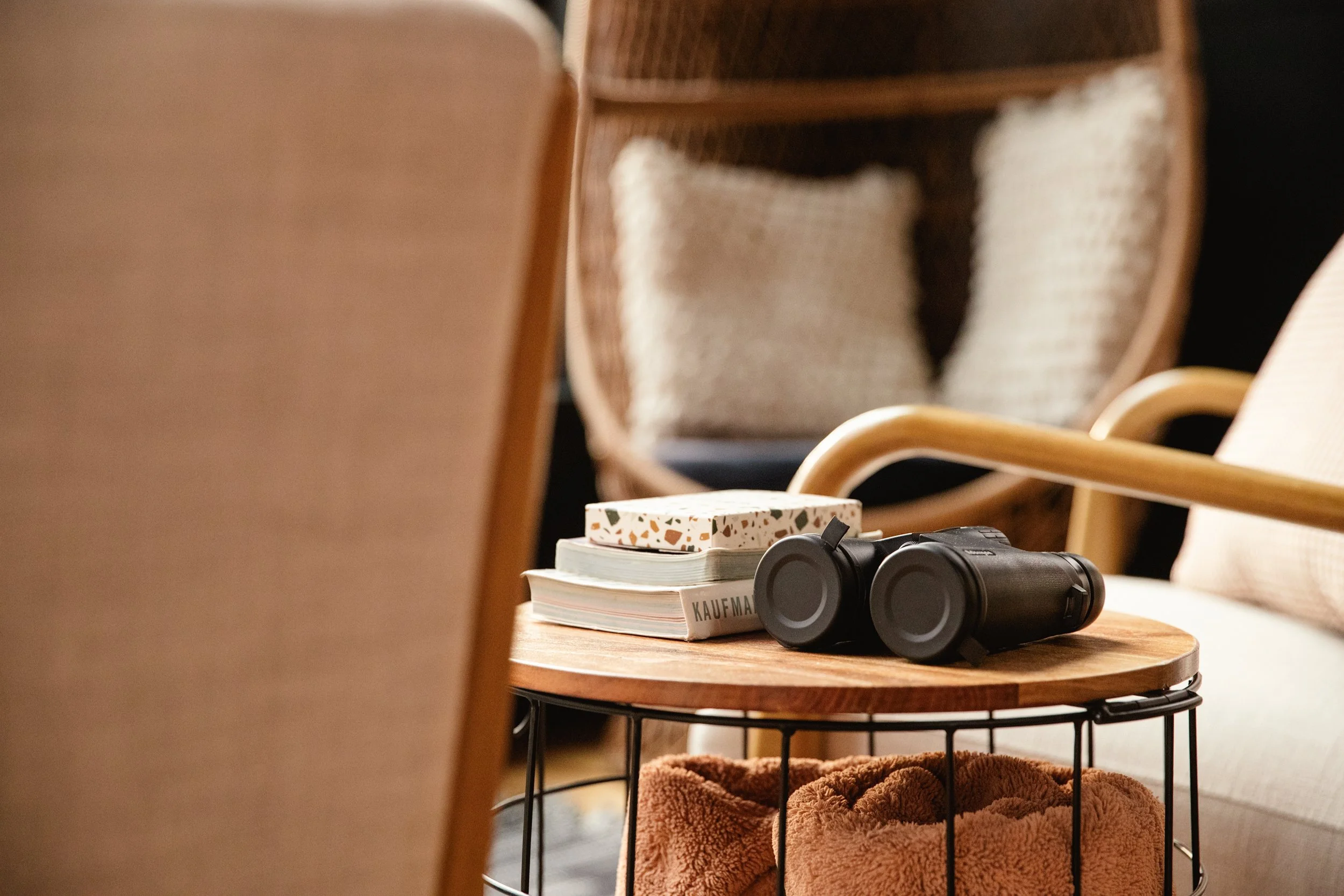 A round wooden coffee table with a black metal wire base, holding a pair of black binoculars, a white book titled 'KAUFMAN', several notebooks, and a folded beige blanket. The background features beige and rattan chairs with light-colored cushions an