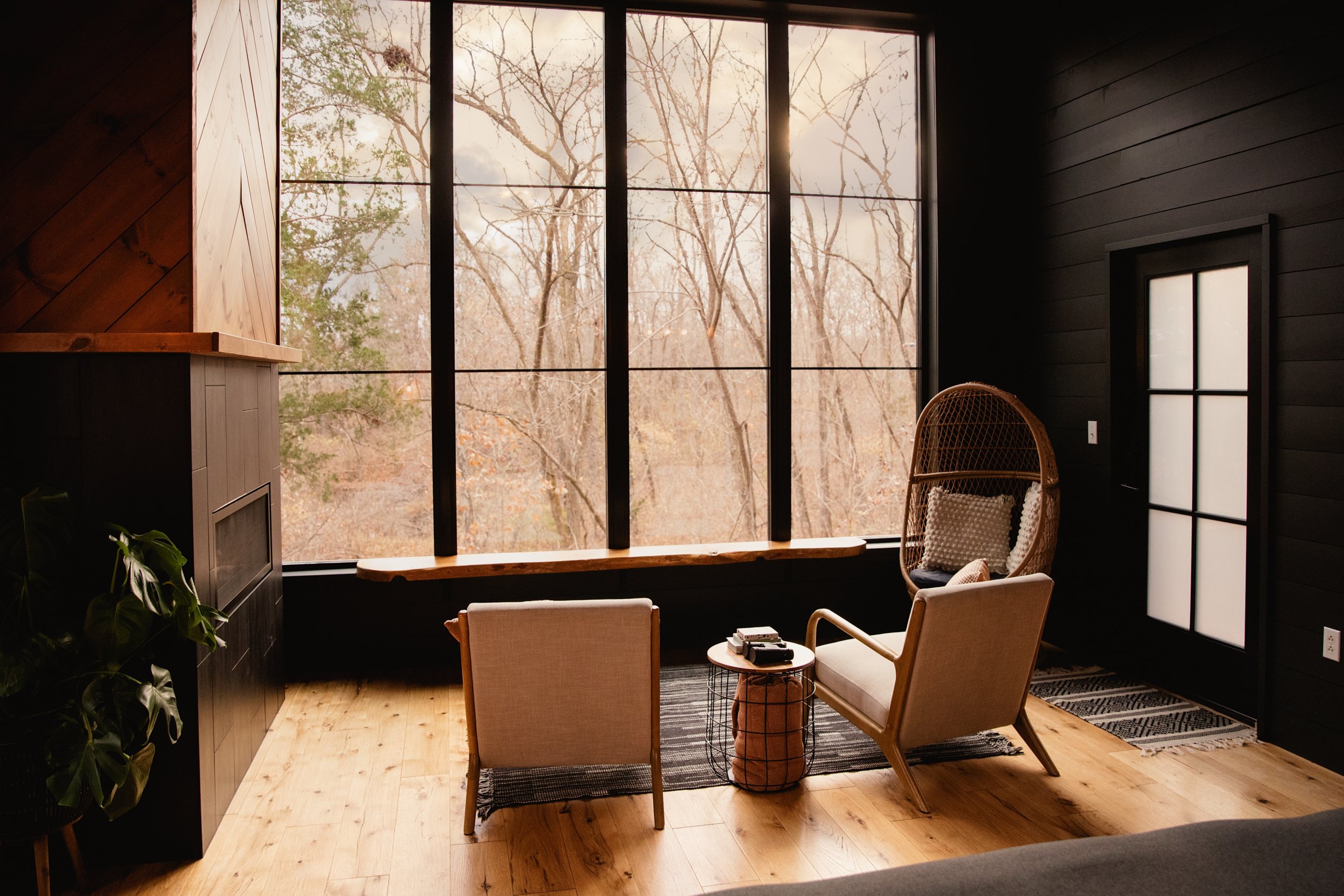 A modern living room with large black-framed window showing trees outside, dark wooden walls, light wooden floor, two beige armchairs, a small round side table with a book and remote, and a wicker chair with cushions.